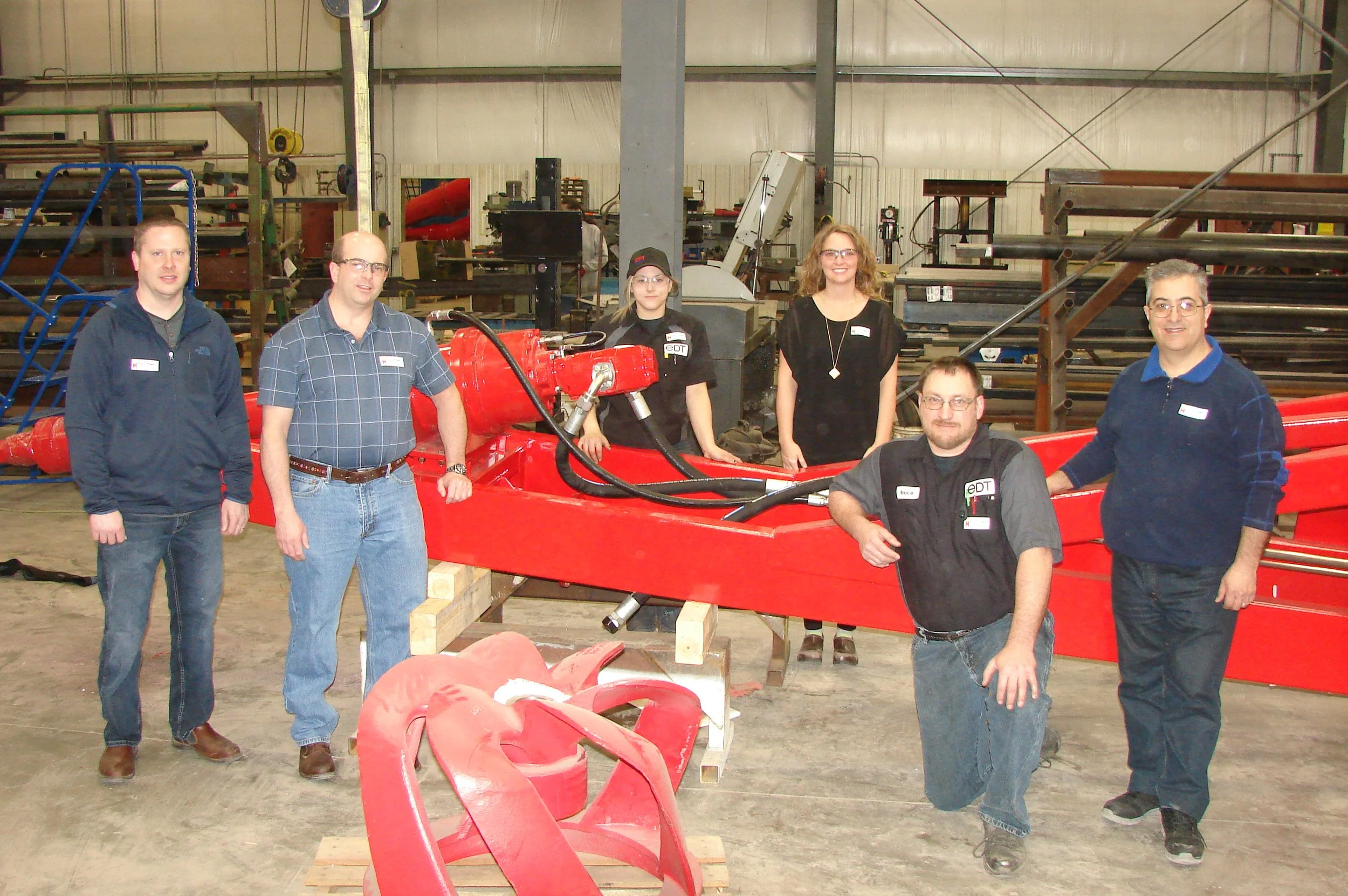 A group of six people, including men and women, standing around a red race car in a workshop or garage. The workshop has various tools, equipment, and metal pipes in the background.