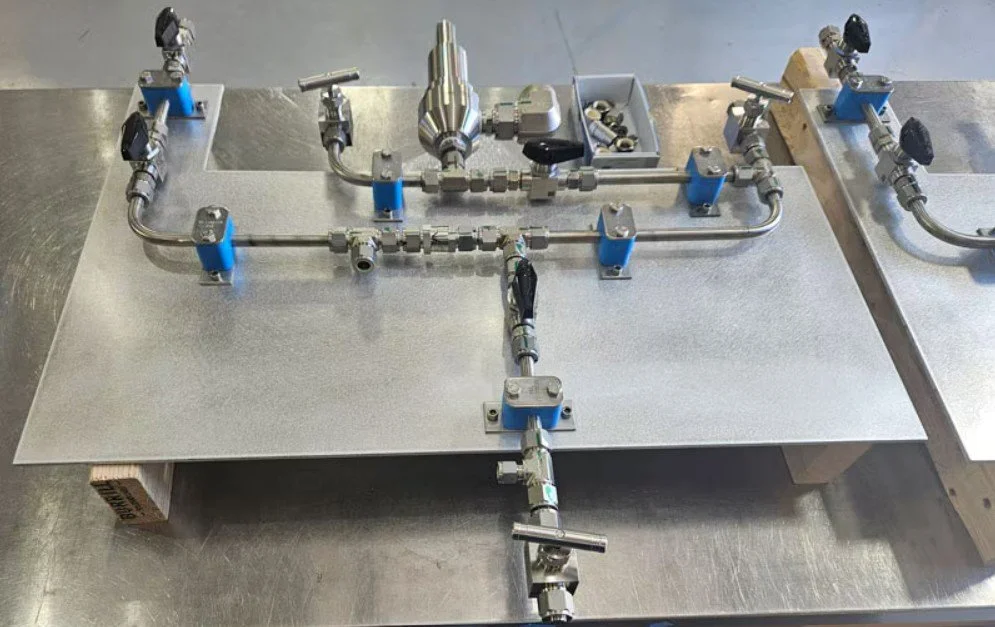 A stainless steel gas delivery system with valves, fittings, and a pressure gauge mounted on a metal plate.