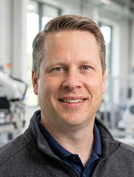 Smiling man in a dark jacket and blue polo shirt, standing in a bright, modern medical or scientific facility.