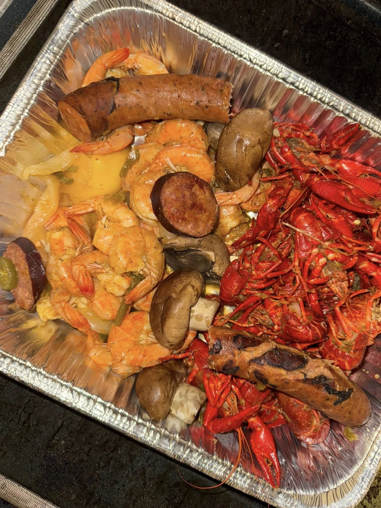 Seafood platter with cooked shrimp, lobster tail, crawfish, smoked sausage, mushrooms, and vegetables in a foil tray.