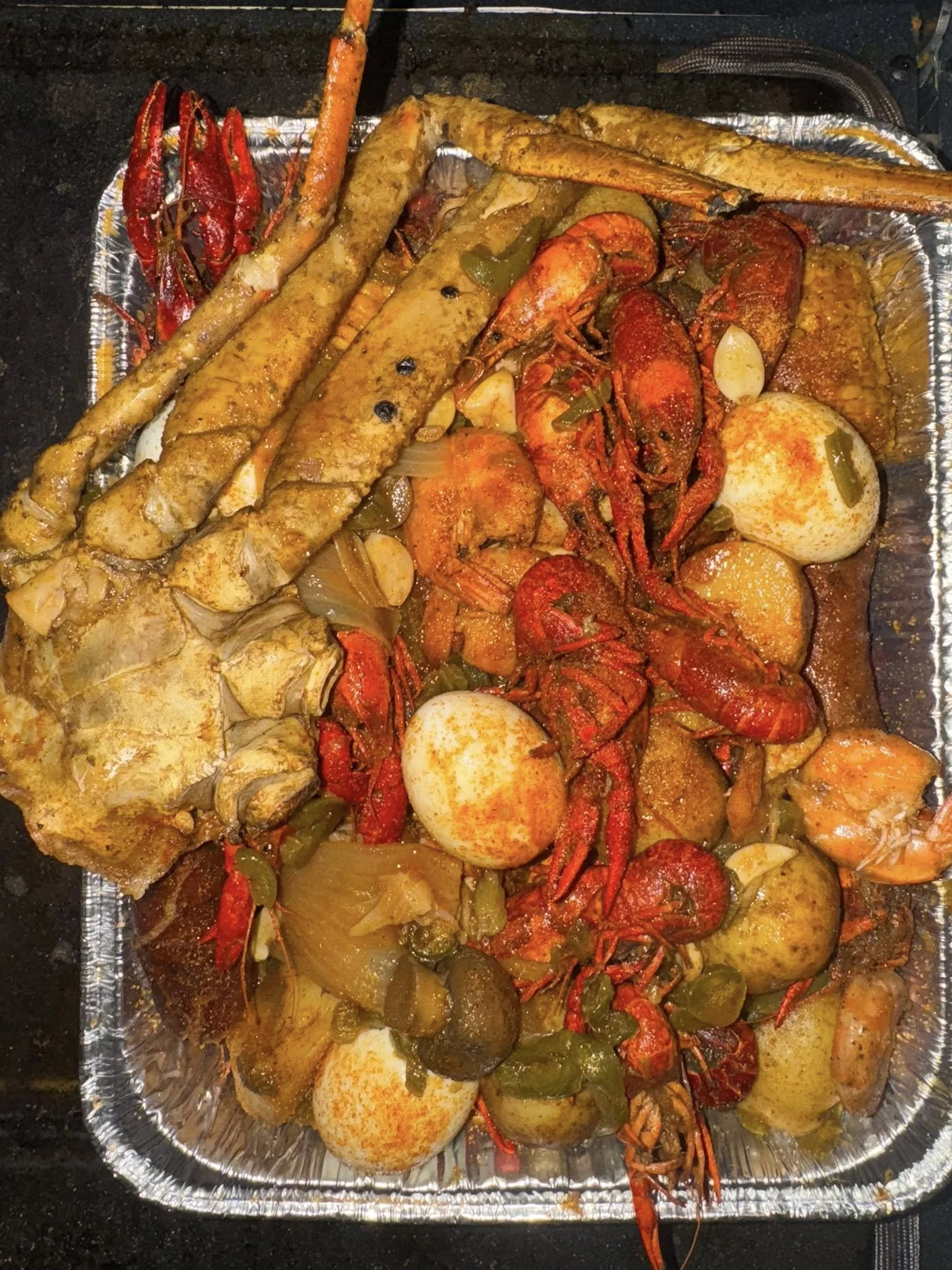 A metal tray filled with seafood stew including crawfish, fish, eggs, potatoes, and vegetables such as peppers and onions, garnished with spices.