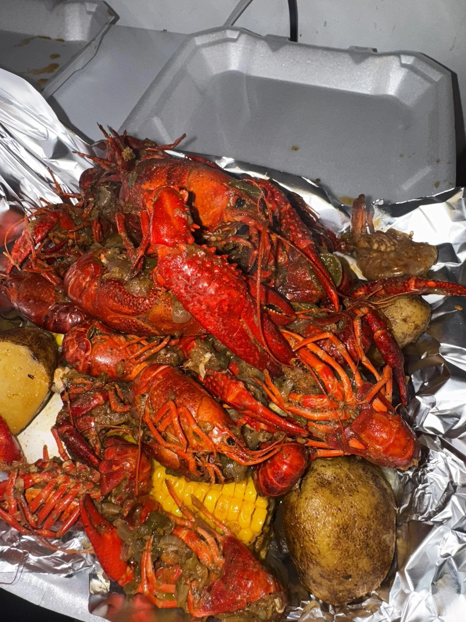 Lobster with melted butter, along with baked potato, corn on the cob, and a piece of bread in foil on a foil-lined surface.
