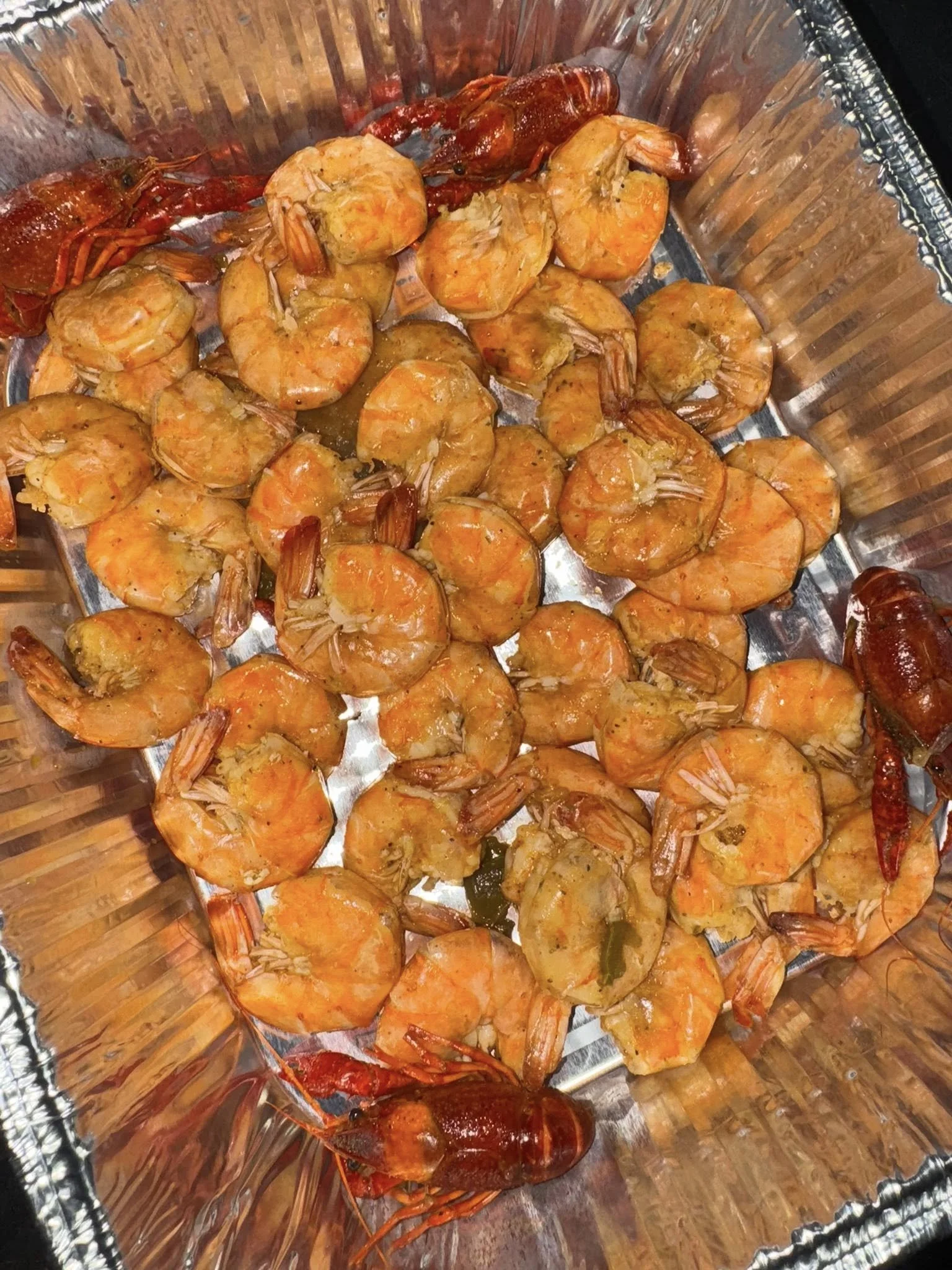 A tray of cooked shrimp with a reddish-orange shell, some with the shell removed, placed on a metallic tray lined with aluminum foil.