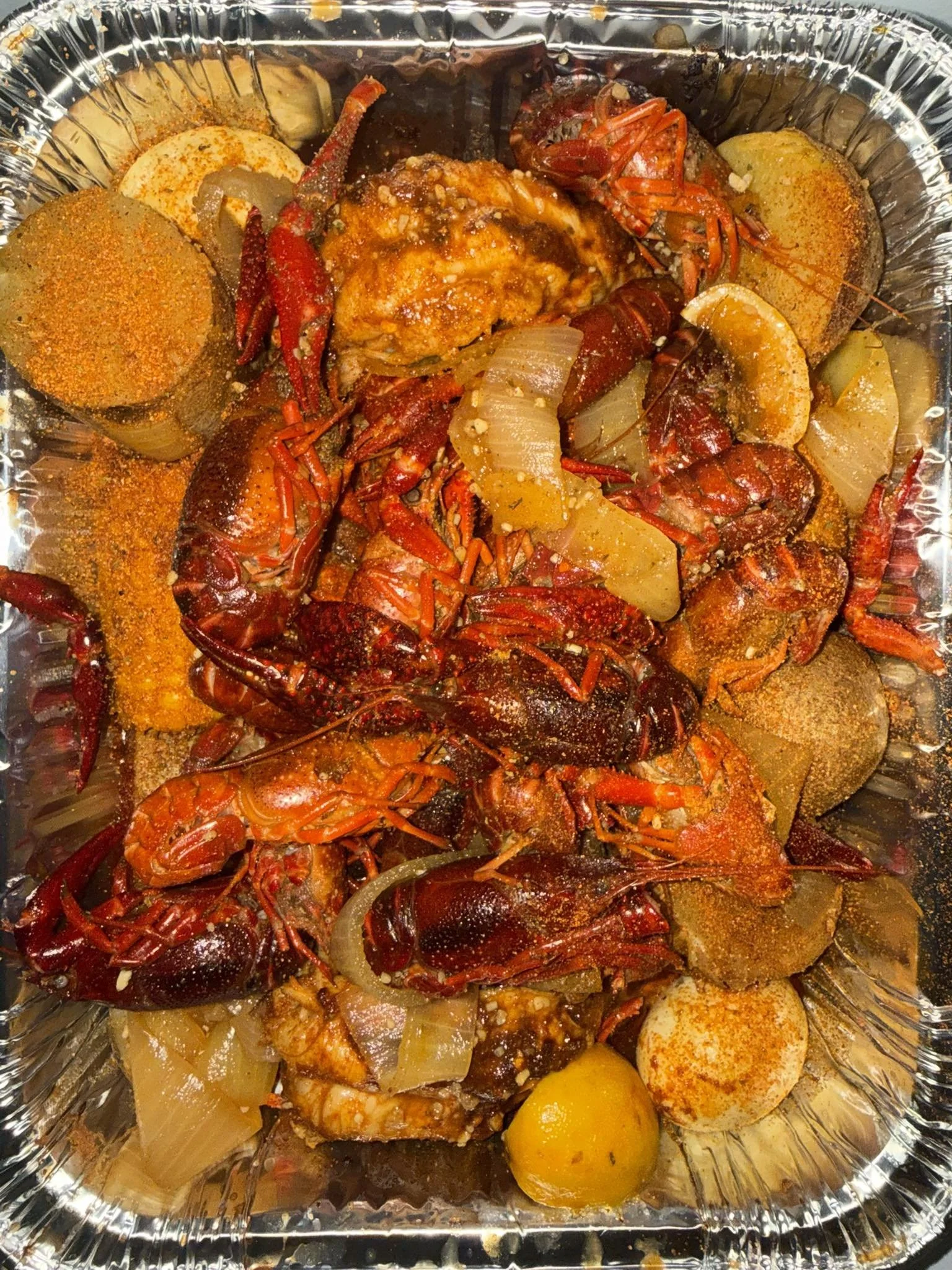 Seafood boil with lobster, crawfish, crab legs, potatoes, corn, and sausage in a foil tray.