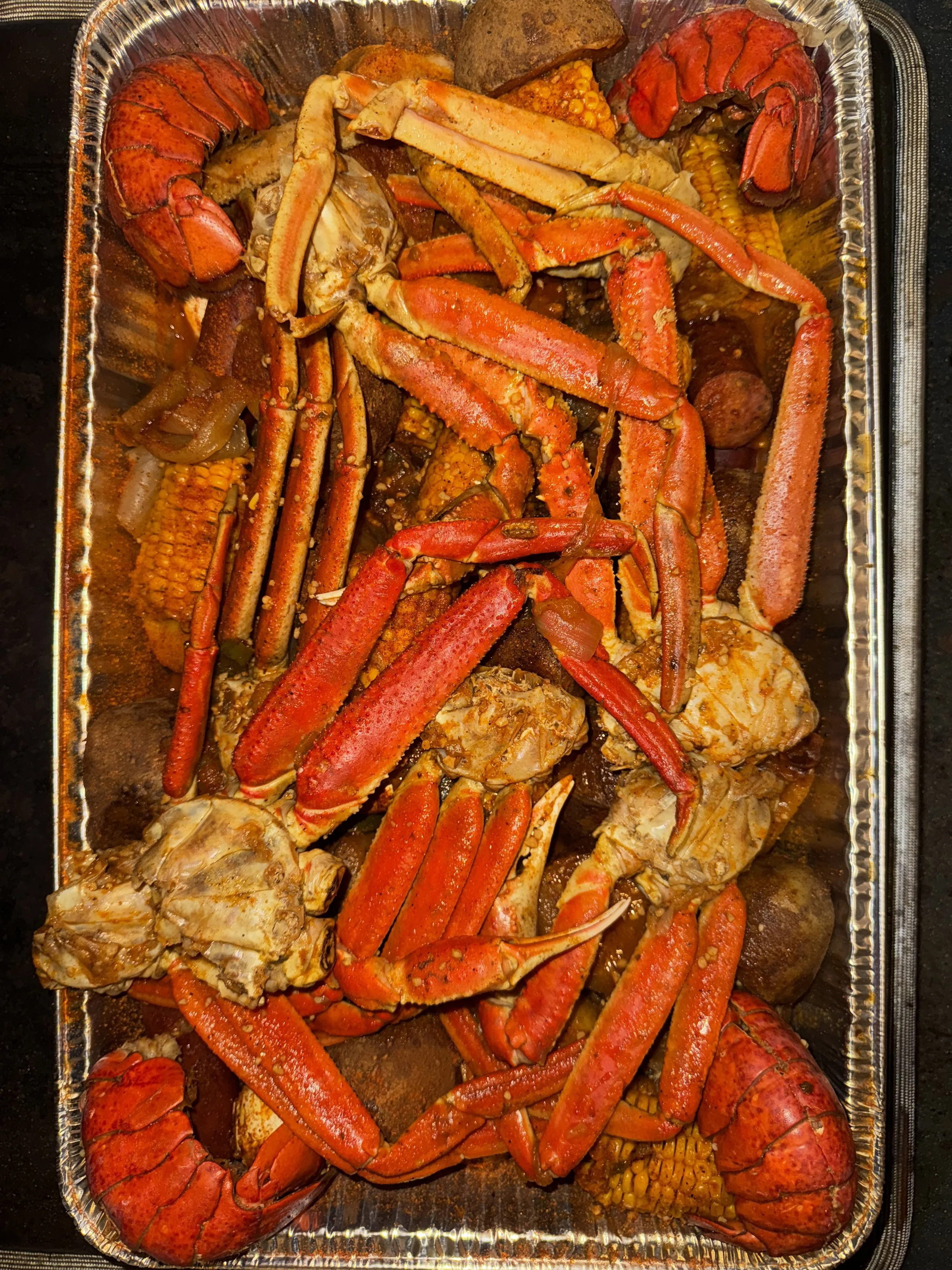 Aluminum tray containing cooked lobster tails, crab legs, and corn on the cob.