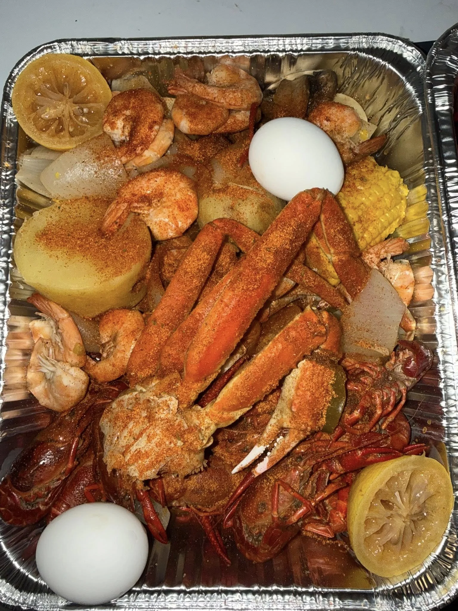Seafood boil with shrimp, lobster, mussels, crab, boiled eggs, lemon slices, corn on the cob, and potatoes in a foil tray