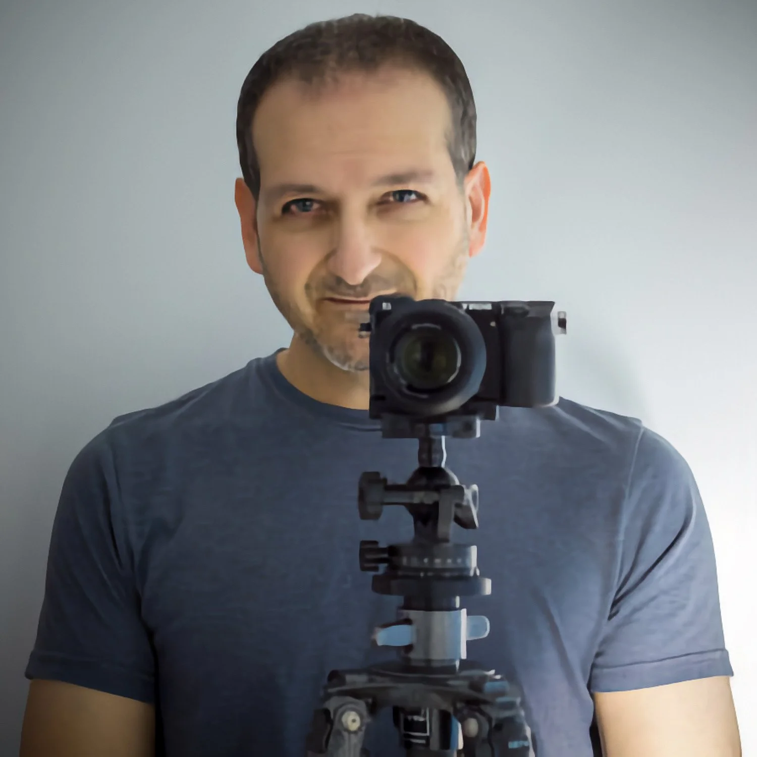 Studio headshot of George Ninos smiling behind a tripod-mounted mirrorless camera against a neutral grey background.