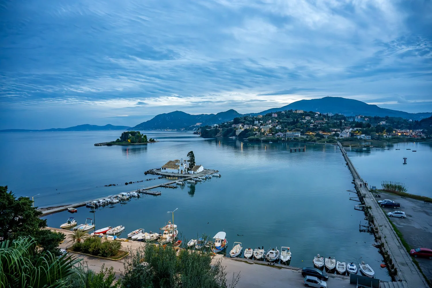Elevated blue-hour view of Kanoni, Corfu: a causeway leading to Vlacherna Monastery and Mouse Island, calm water and distant hills.