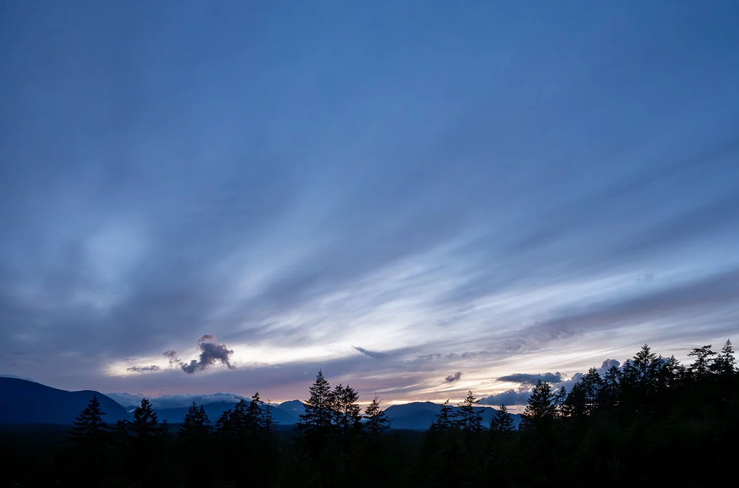Wind-brushed clouds streaking across a deep blue twilight sky above a silhouetted mountain ridgeline and treeline, Port Alberni.