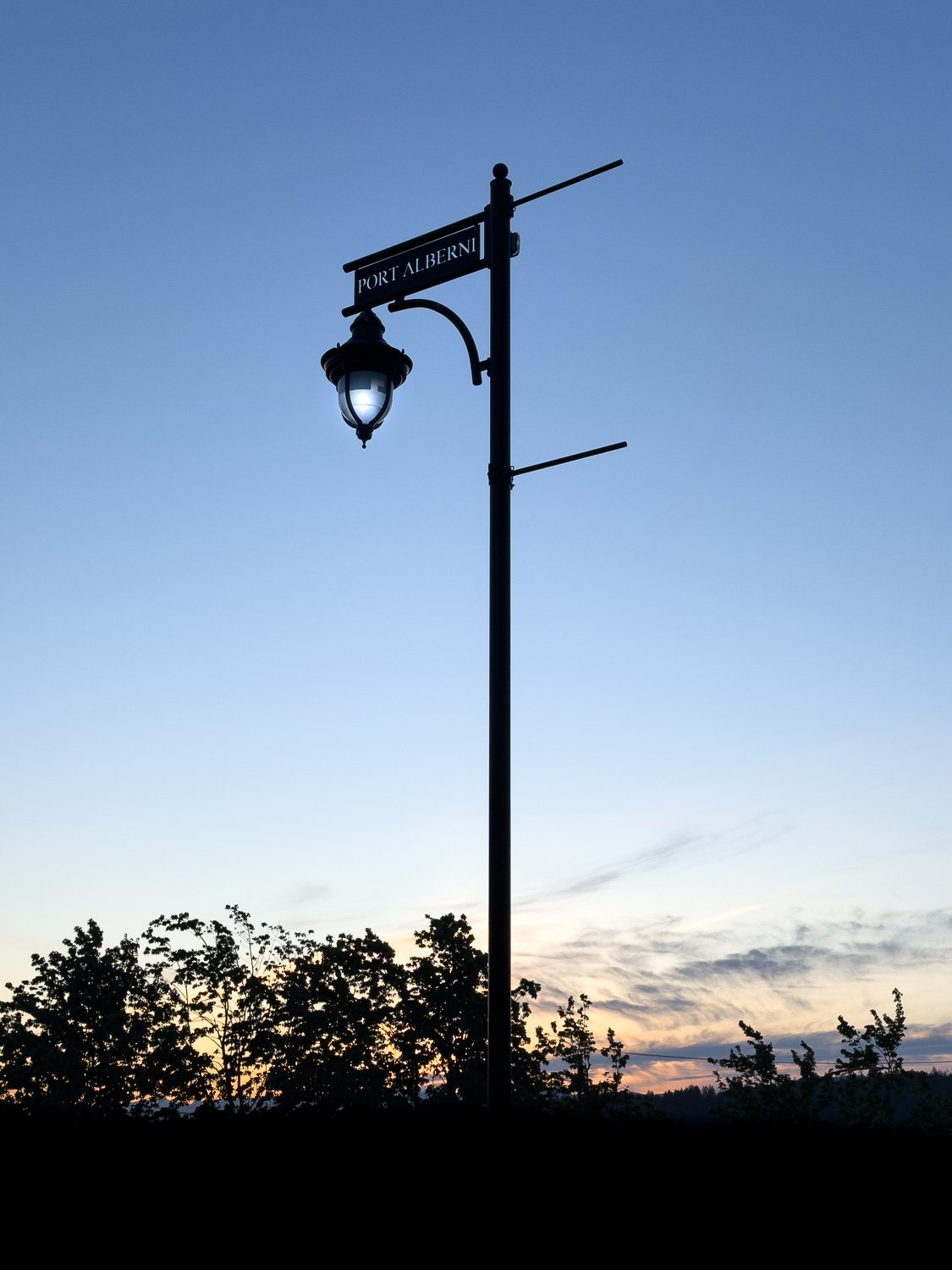 Port Alberni heritage street lamp silhouetted against a blue and amber twilight sky over a dark treeline on Vancouver Island.