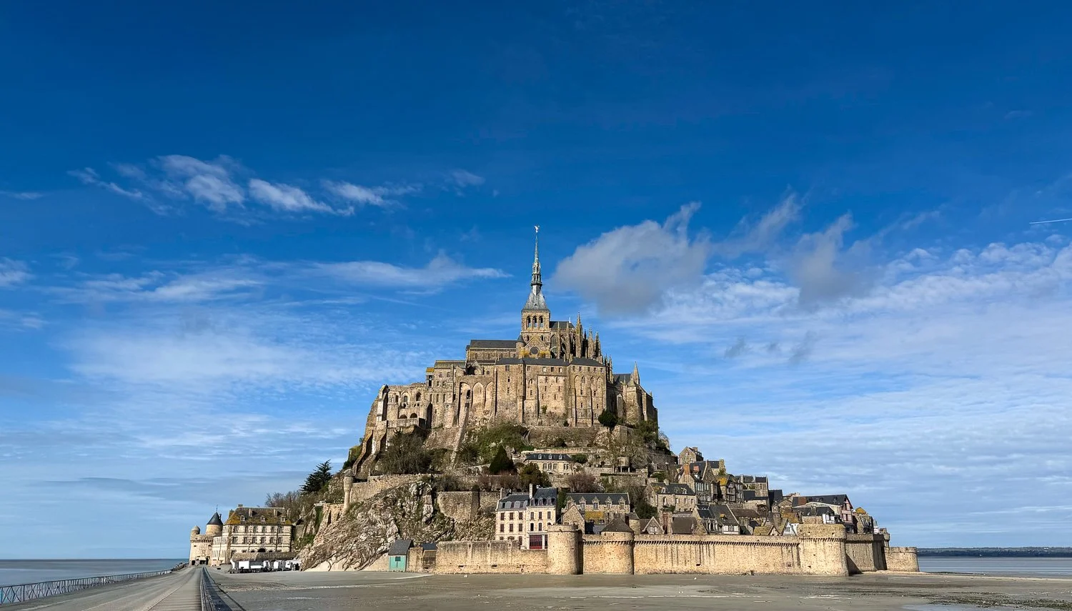 The island abbey of Mont Saint-Michel rising from low-tide sand flats under a blue sky with scattered cloud, the causeway entering at left.