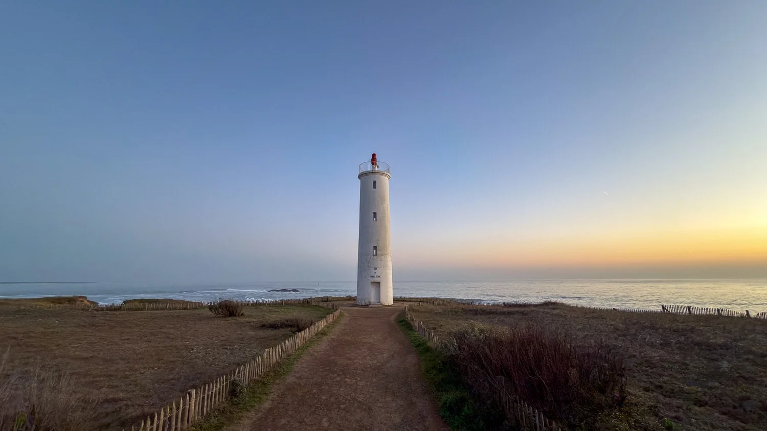 A white lighthouse at the end of a path on the French Atlantic coast, soft pastel sunset sky and ocean behind.