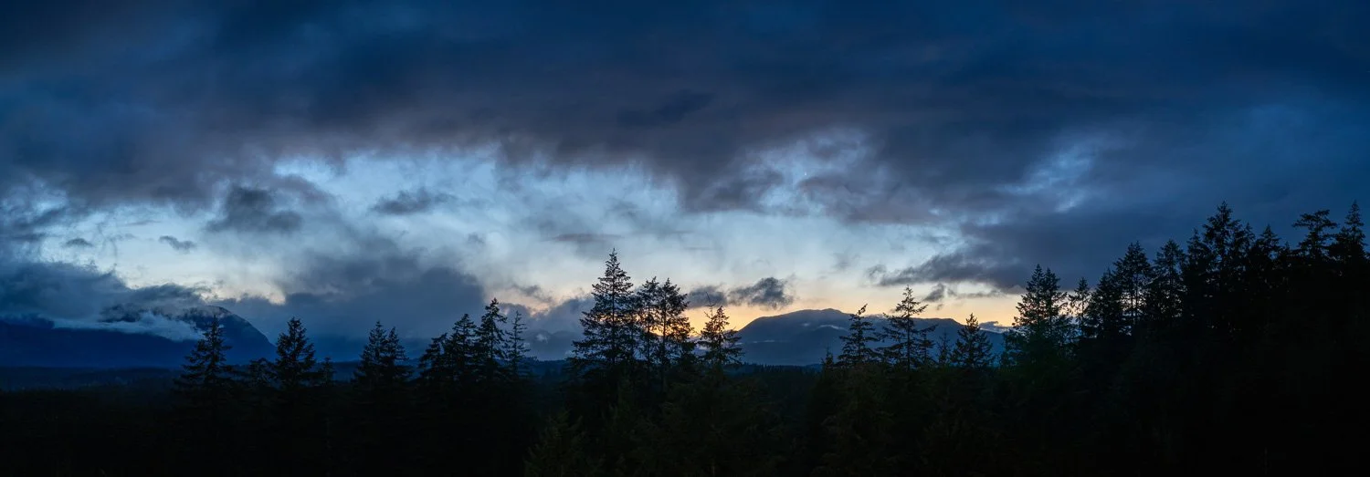 Heavy blue-grey clouds over a silhouetted mountain ridgeline and conifer treeline, with a seam of gold sunset light breaking through low on the horizon, Port Alberni.