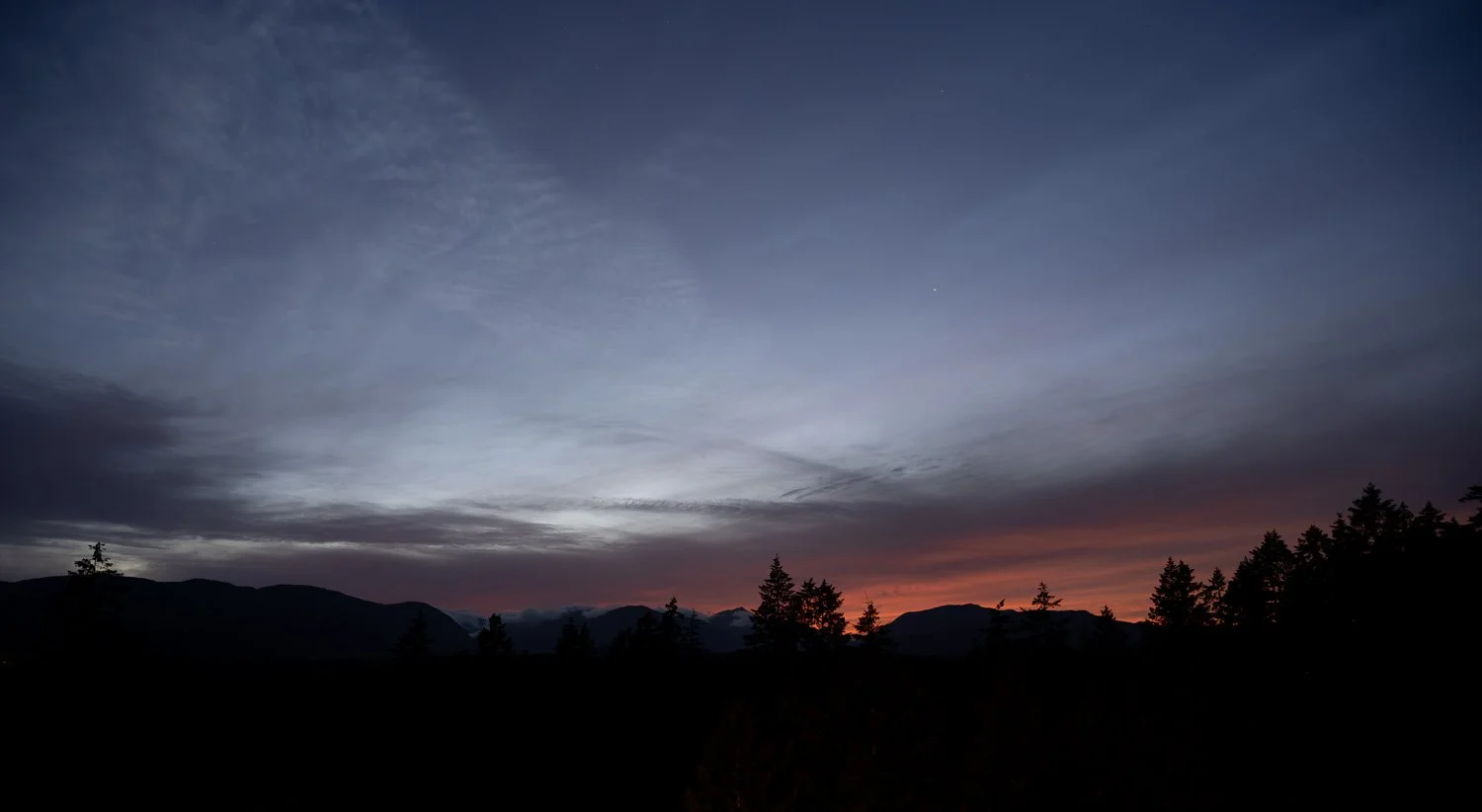 A thin band of ember-orange light along the horizon beneath a deep blue twilight sky, with a silhouetted ridgeline and firs, Port Alberni.