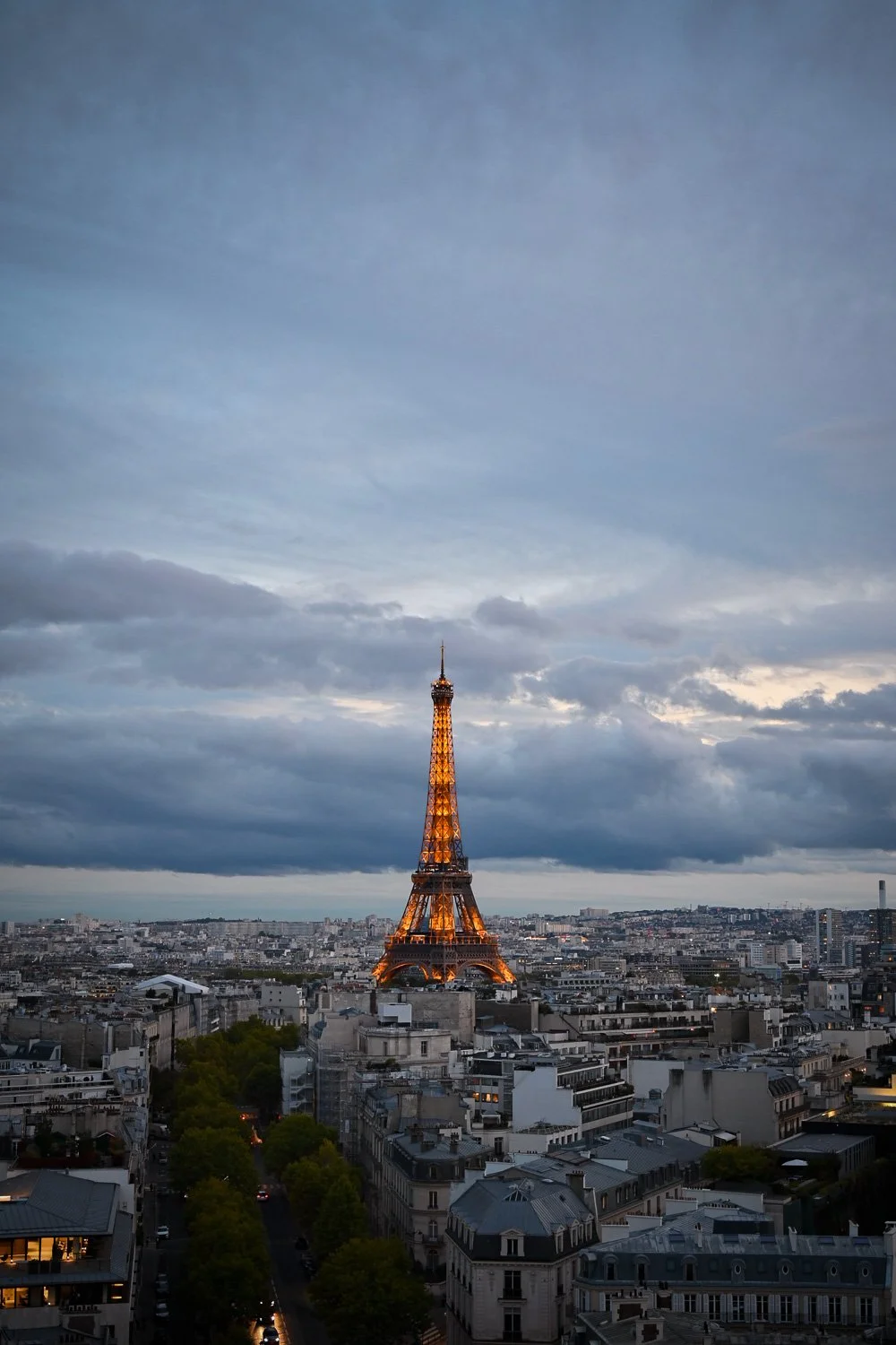 The illuminated Eiffel Tower at dusk rising above the Paris cityscape under a cloudy evening sky, vertical composition.