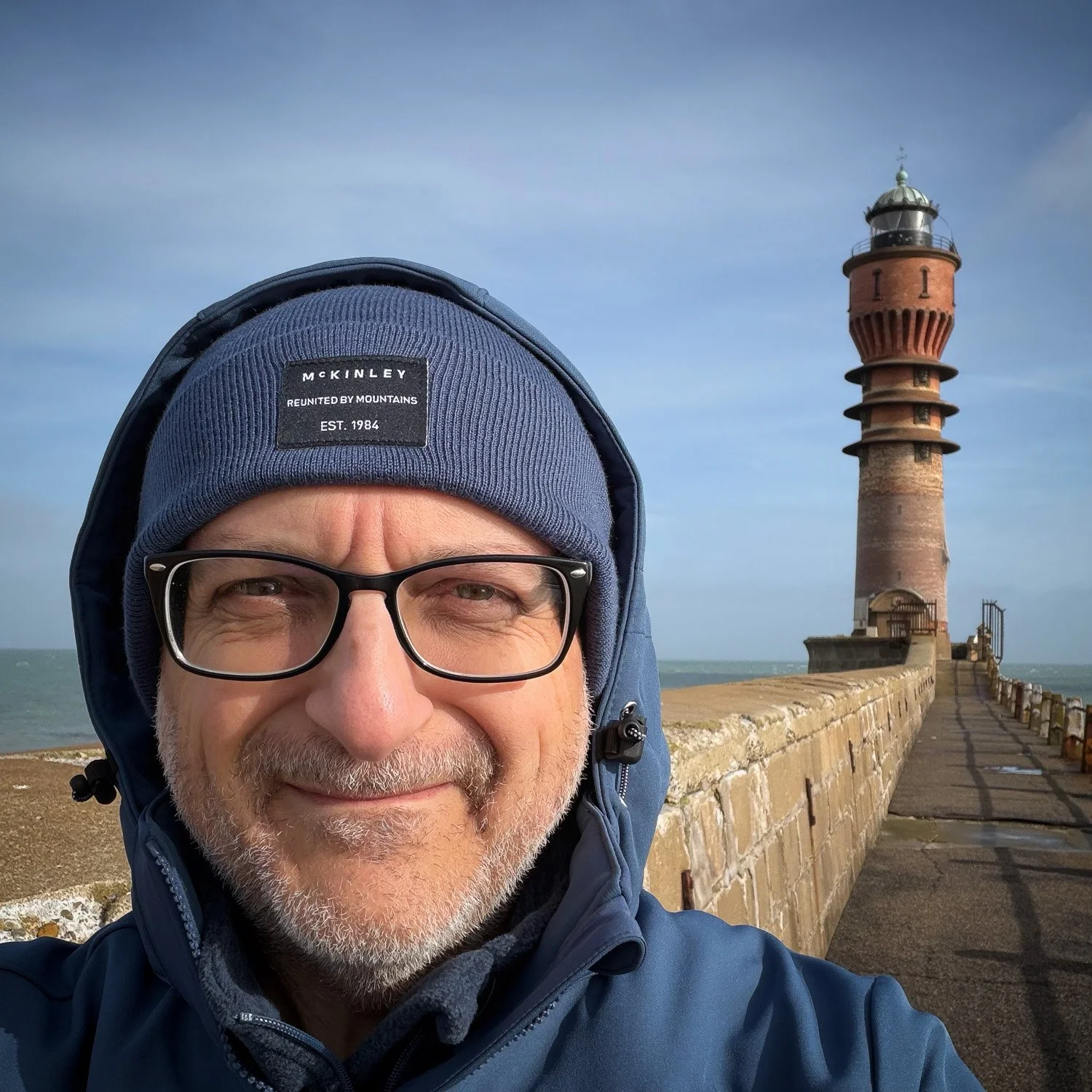 George Ninos in a hooded jacket and beanie smiling on the Dunkerque pier, the brick lighthouse behind under a blue sky.