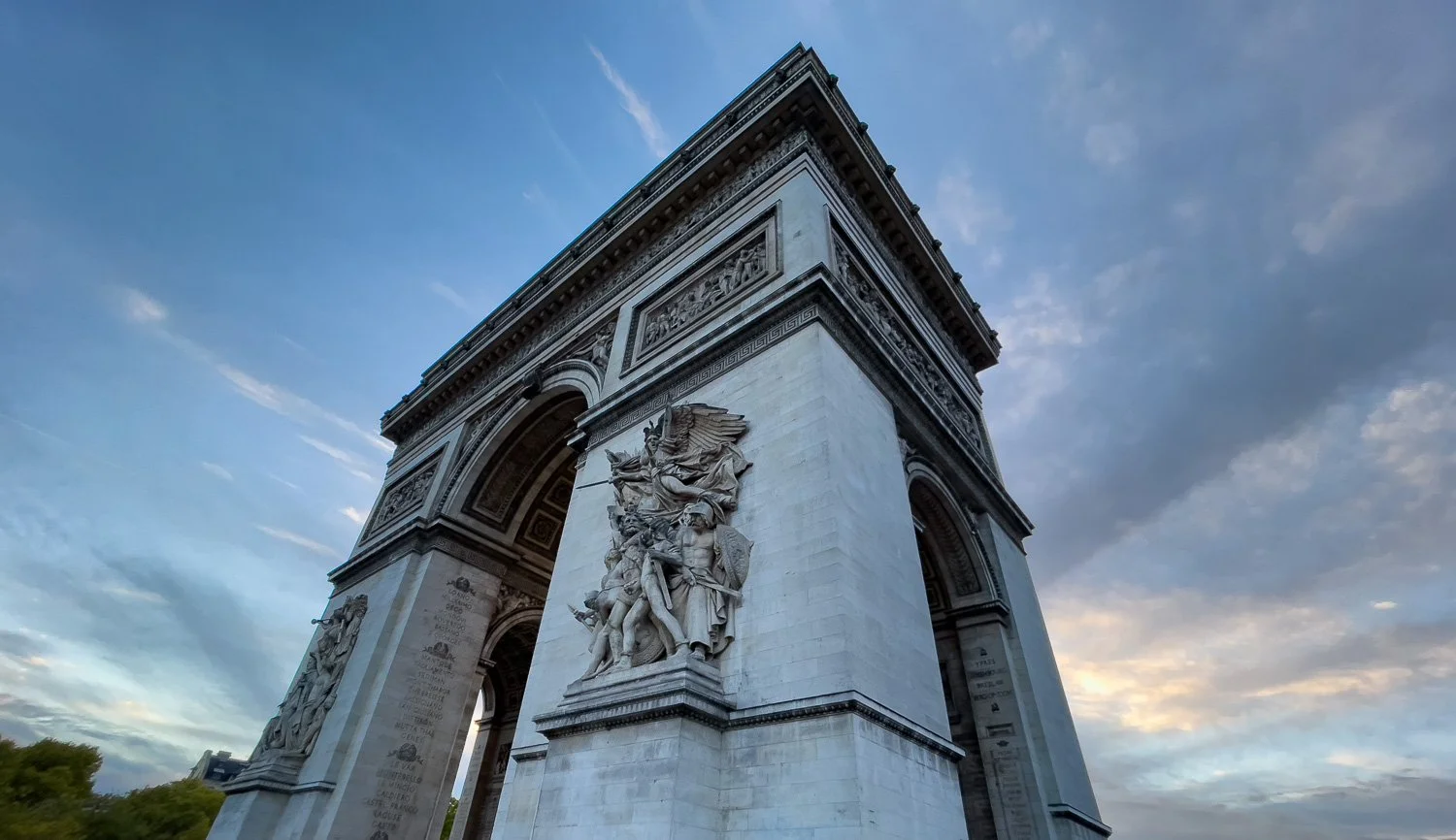 Dramatic upward view of the Arc de Triomphe in Paris against a dusk sky with streaking clouds, sculptural detail prominent.