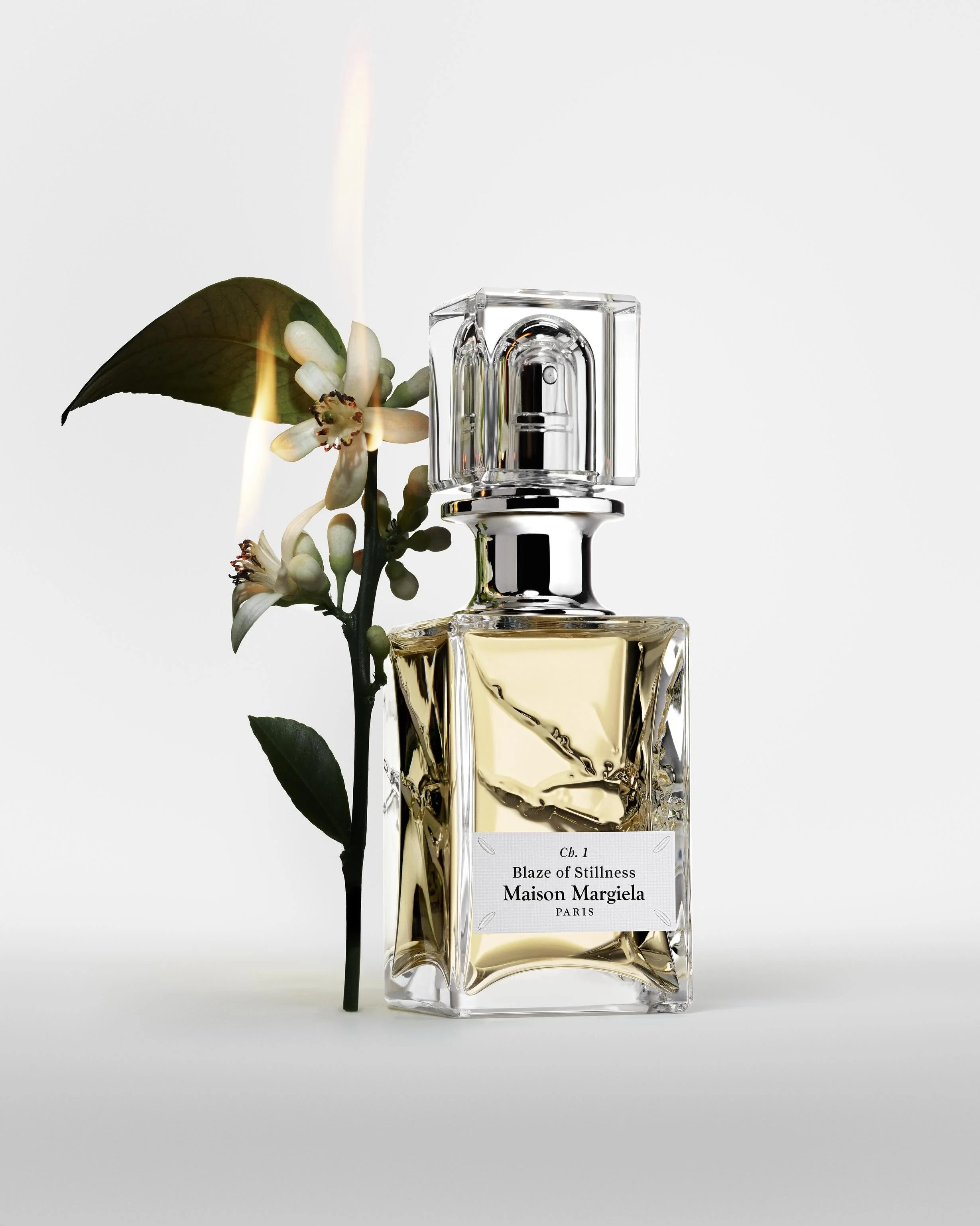 A perfume bottle labeled 'Ch. 1 Blaze of Stillness Maison Margiela Paris' with a small branch of white flowers and green leaves beside it