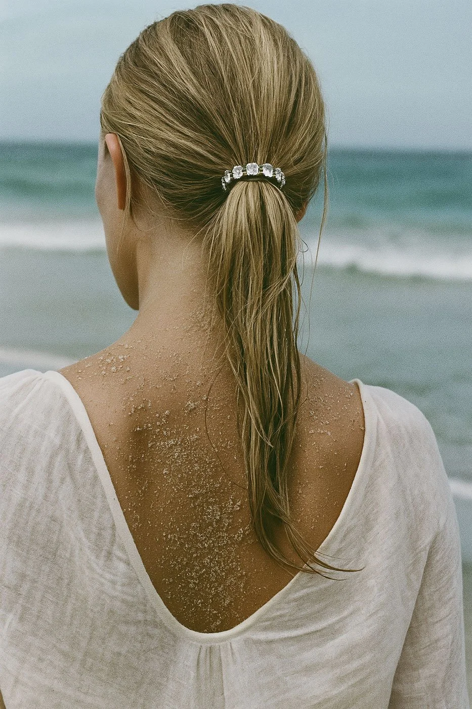 Back of a woman with wet hair tied in a ponytail, wearing a white top, standing on a beach with sand on her shoulders and neck, facing the ocean.