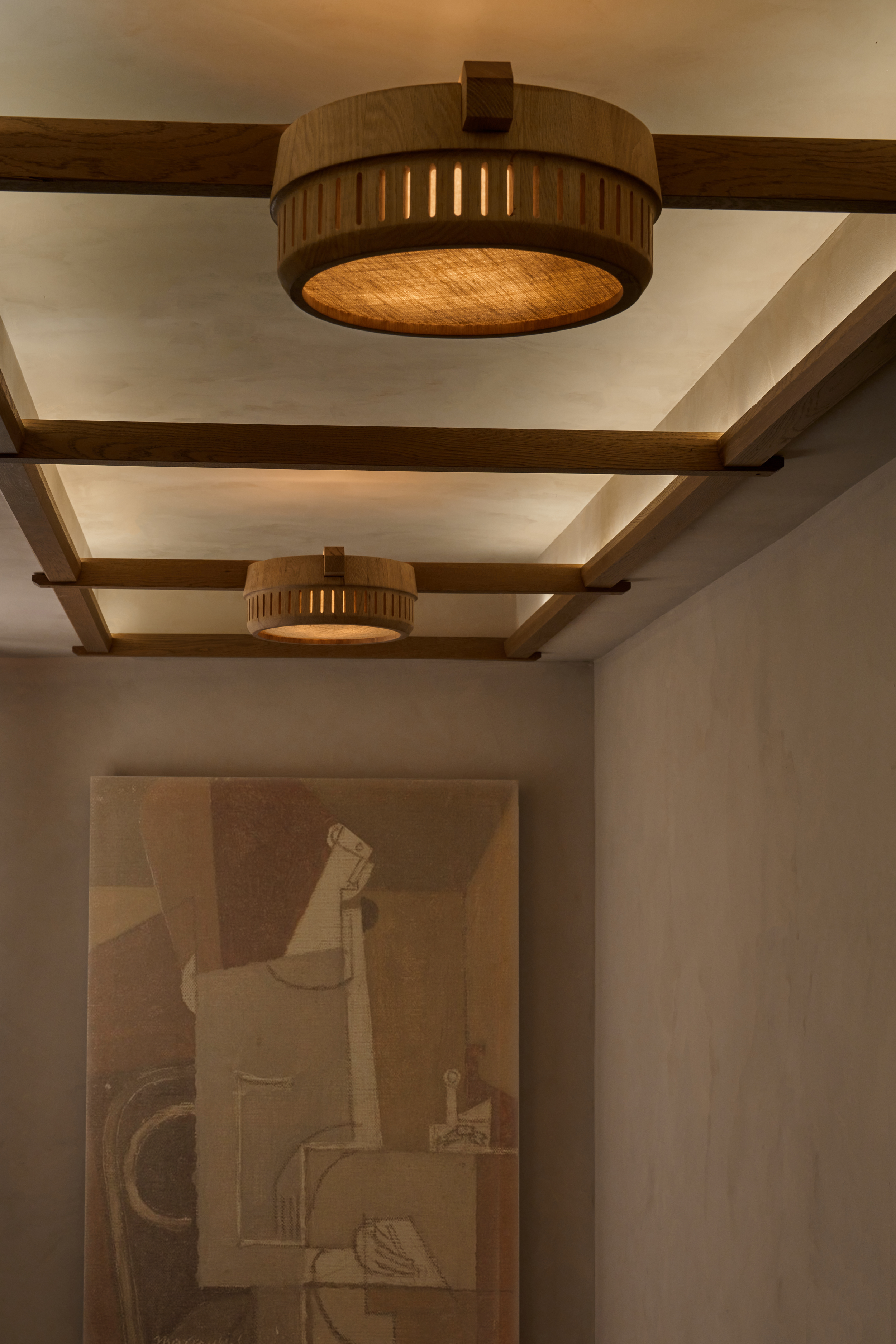 Wooden ceiling light fixtures with slatted design hanging from wooden beams in a room with beige walls. Partially visible abstract artwork on the wall.