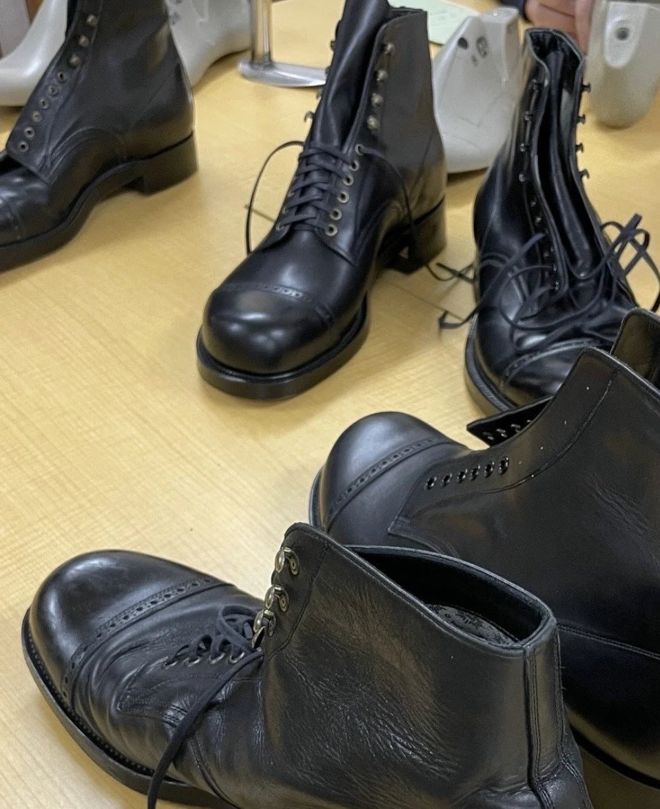Black leather boots, many with laces, placed on a wooden floor.