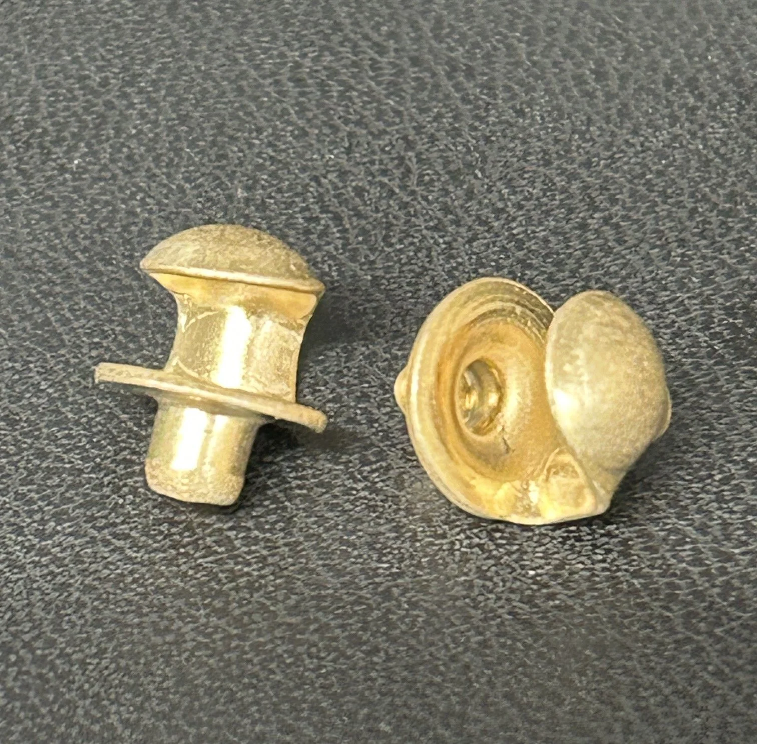 Two gold-colored, short, round, decorative metal pieces with hollow centers and flat bases on a black textured surface.