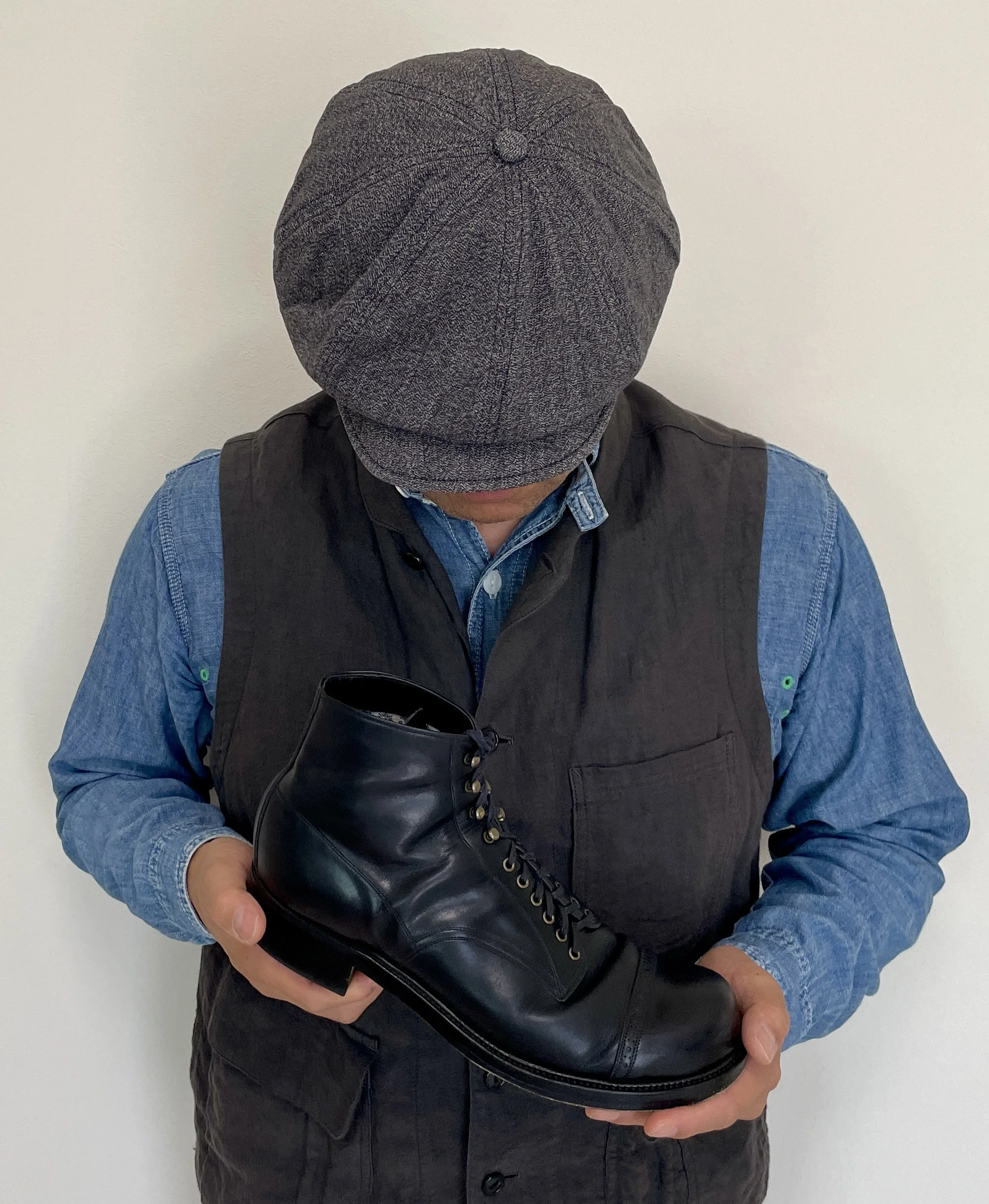 Person wearing a gray knit cap, a black vest over a denim shirt, holding a black leather boot in front of a plain light-colored wall.
