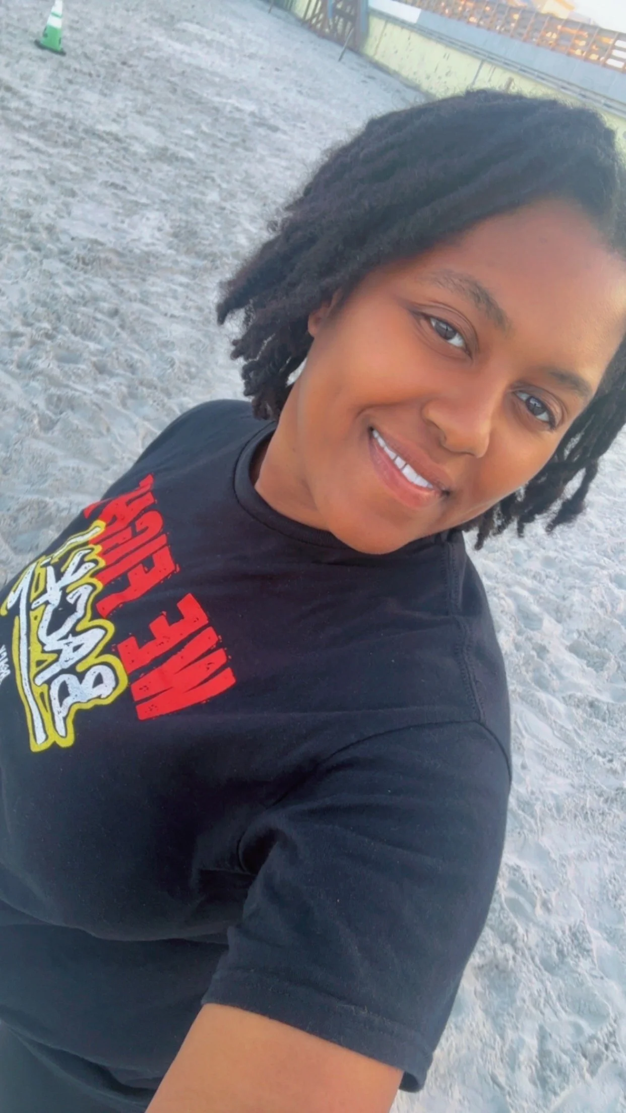A young woman taking a selfie at the beach with sand and wooden fencing in the background. She is wearing a black T-shirt with colorful graphics.