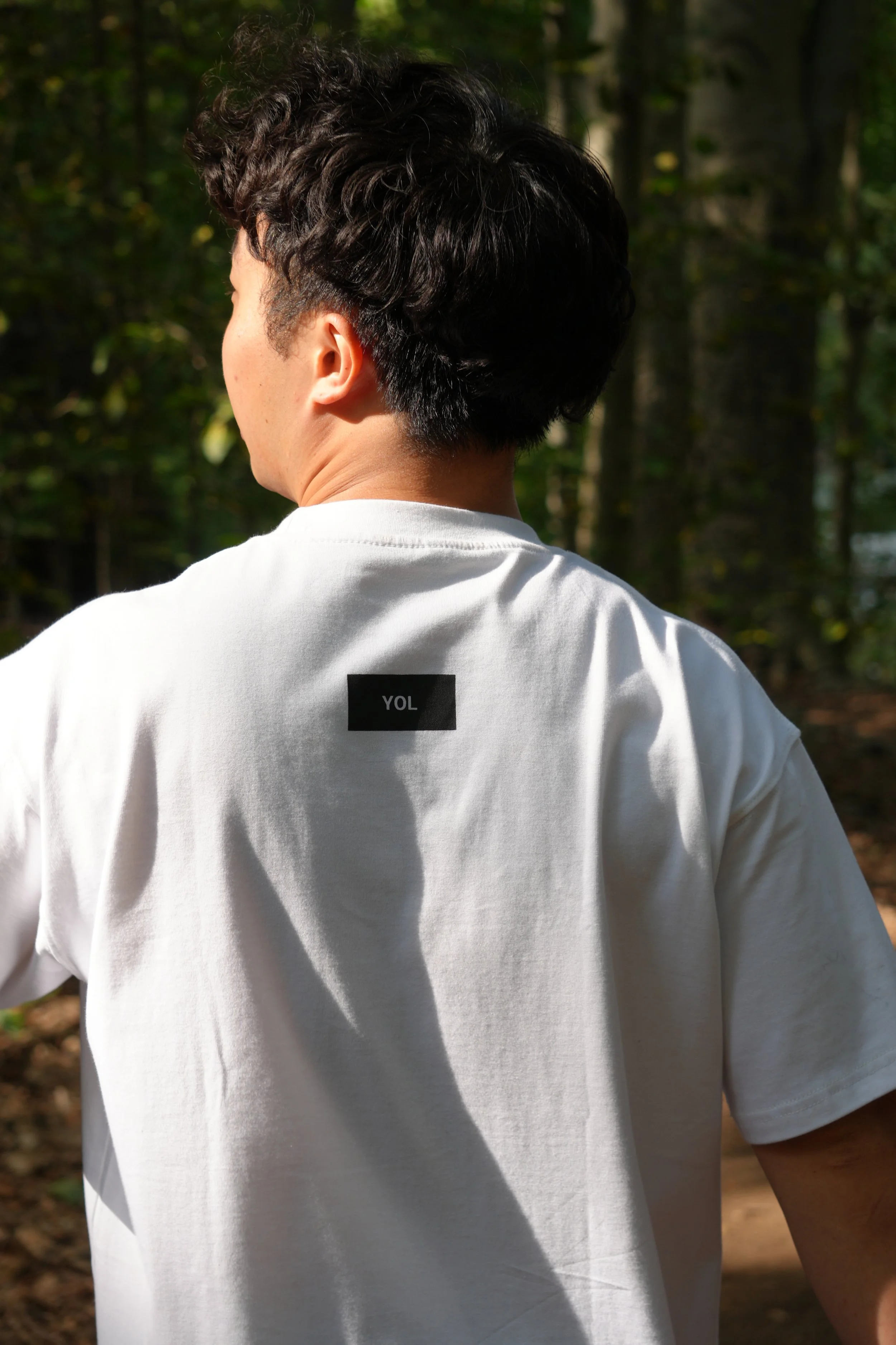 Back of a person with dark, curly hair wearing a white t-shirt with a black label that reads 'YOL' on the back. The person is outdoors in a forested area with green trees in the background.