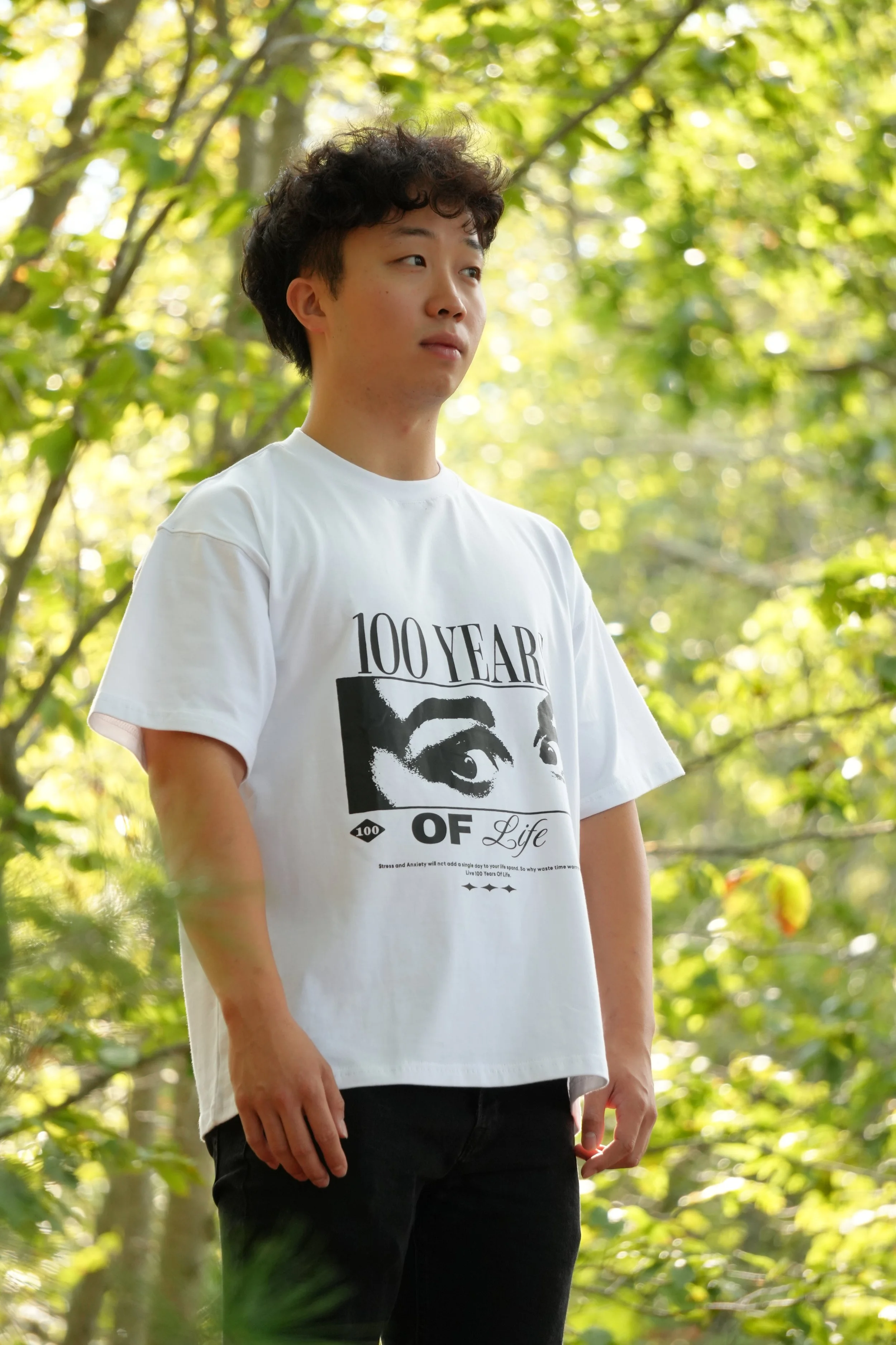 A young man with curly dark hair wearing a white T-shirt and black pants standing outdoors in a green, leafy forested area.