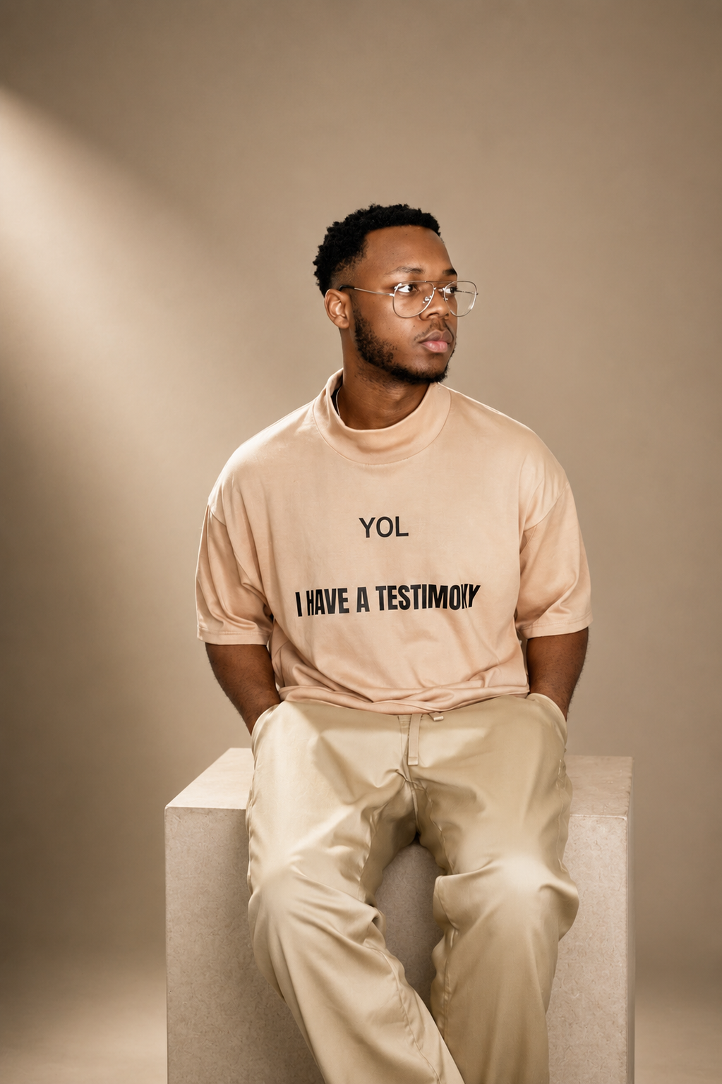A young man with glasses and a beard sitting on a beige cube, wearing a light beige t-shirt with text and beige pants, looking to the side against a neutral background.
