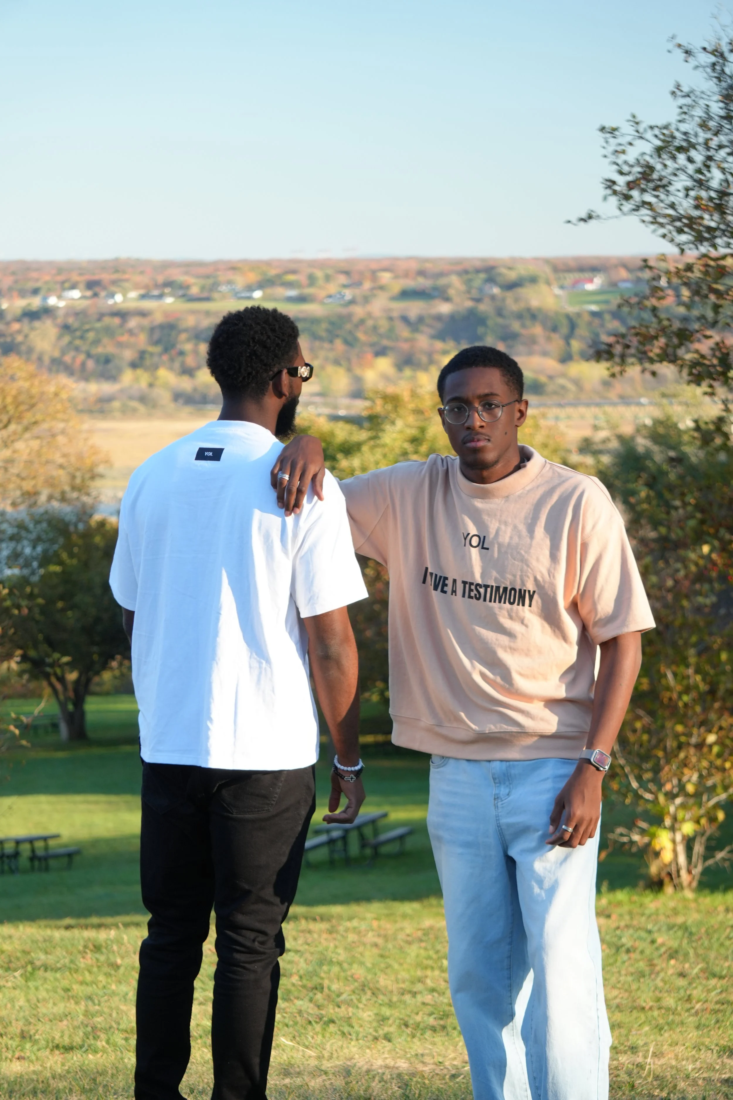 Two young men standing outdoors in a park with rolling hills and houses in the background. One man faces away, wearing a white T-shirt, and the other faces forward, wearing a beige T-shirt with text, glasses, and a watch.