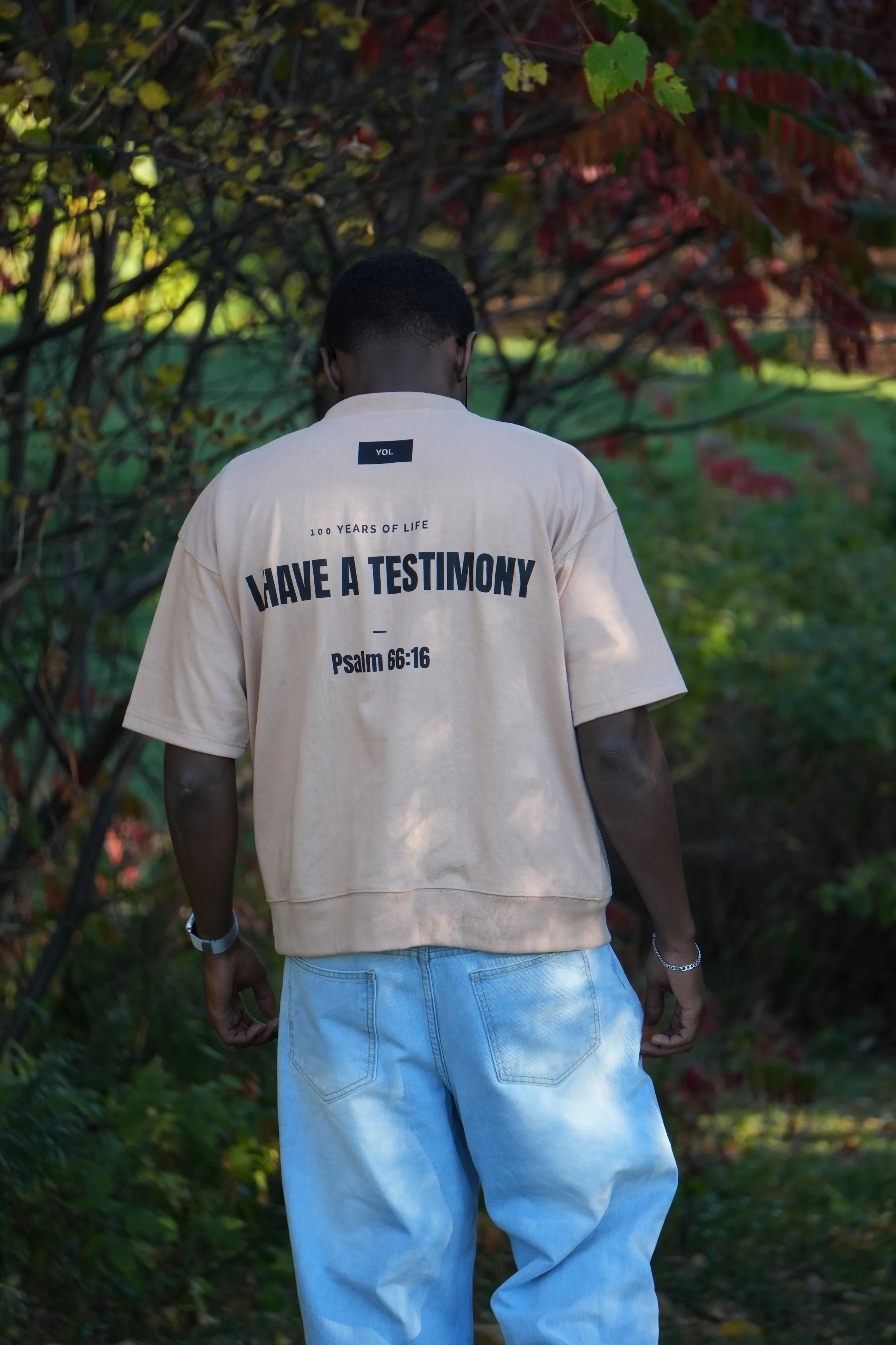 A person with dark skin, short black hair, wearing a beige t-shirt with religious text and jeans, standing outdoors among trees with green and red leaves, facing away from the camera.