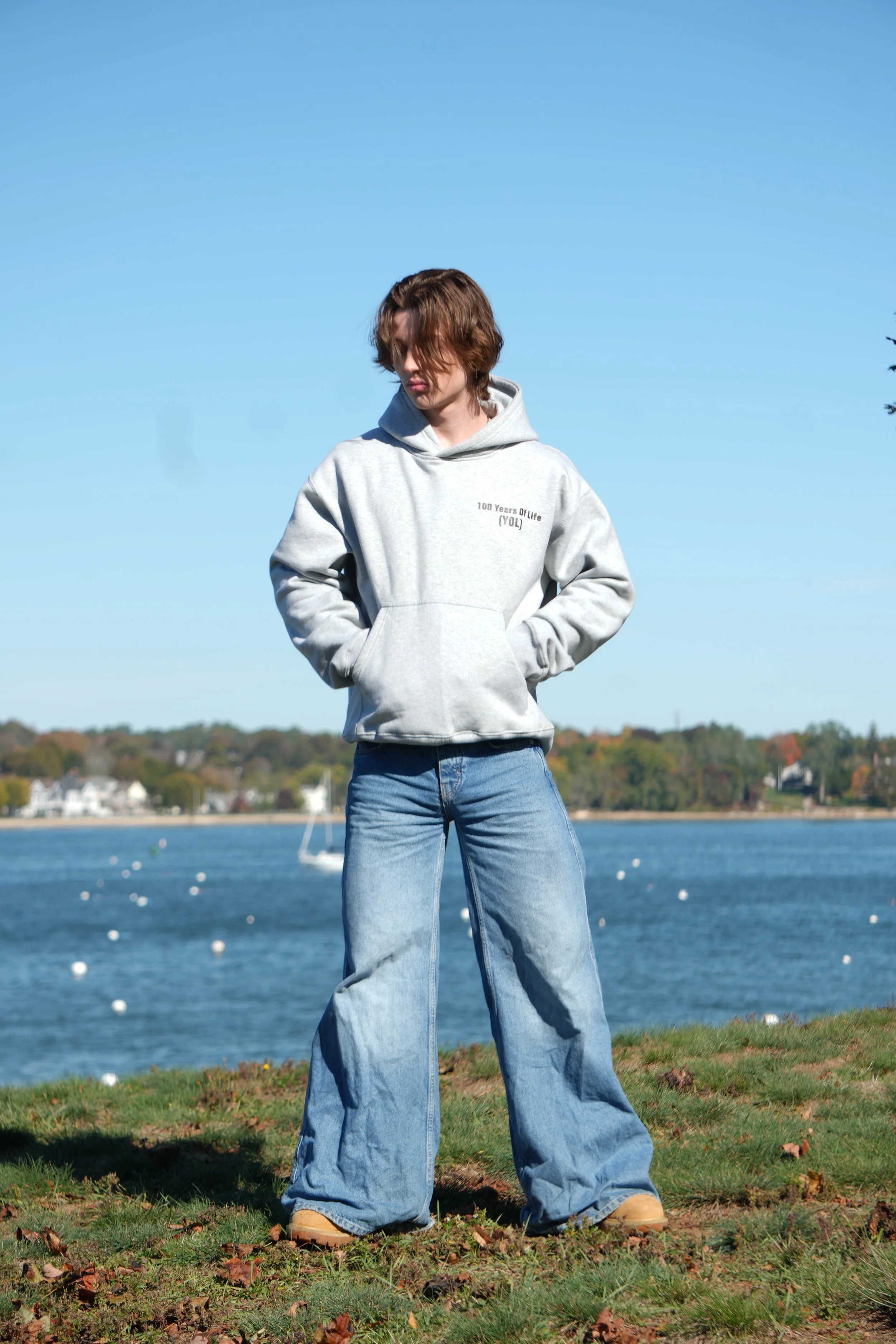 A person with shoulder-length brown hair standing outdoors near a body of water, wearing a light gray hoodie with text on it, blue wide-leg jeans, and beige shoes, with trees and houses in the background under a clear blue sky.