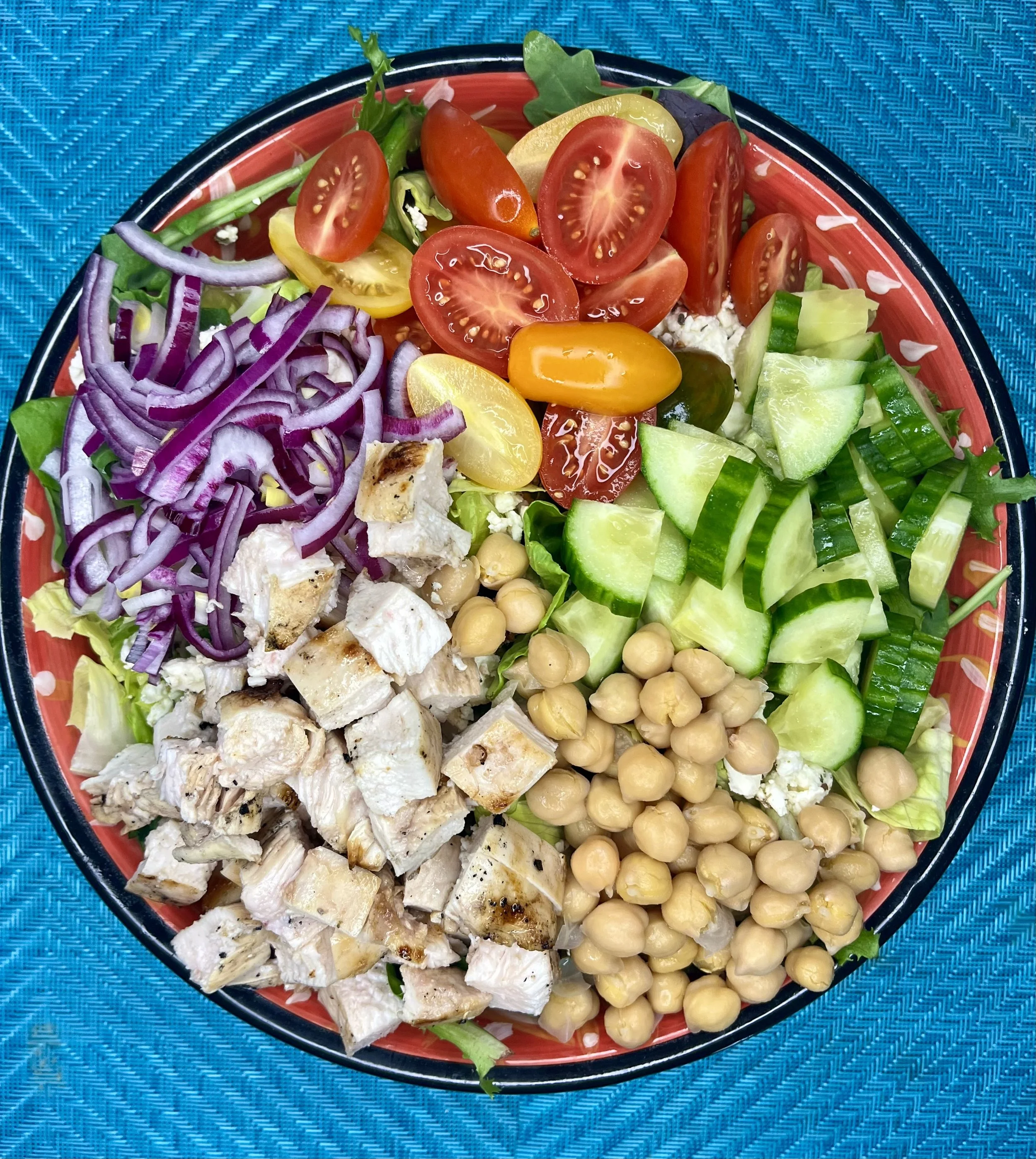 A bowl containing a salad with cherry tomatoes, cucumbers, red onion, chickpeas, and chopped chicken on a blue textured surface.