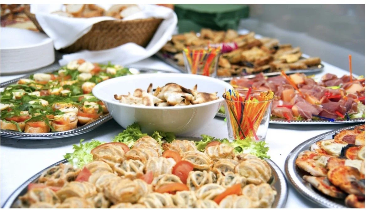 Assorted appetizers and finger foods on a table, including sushi rolls, sliced cheese with toppings, mushrooms, cured meats, and pizza, with some colorful straws and a basket of bread in the background.