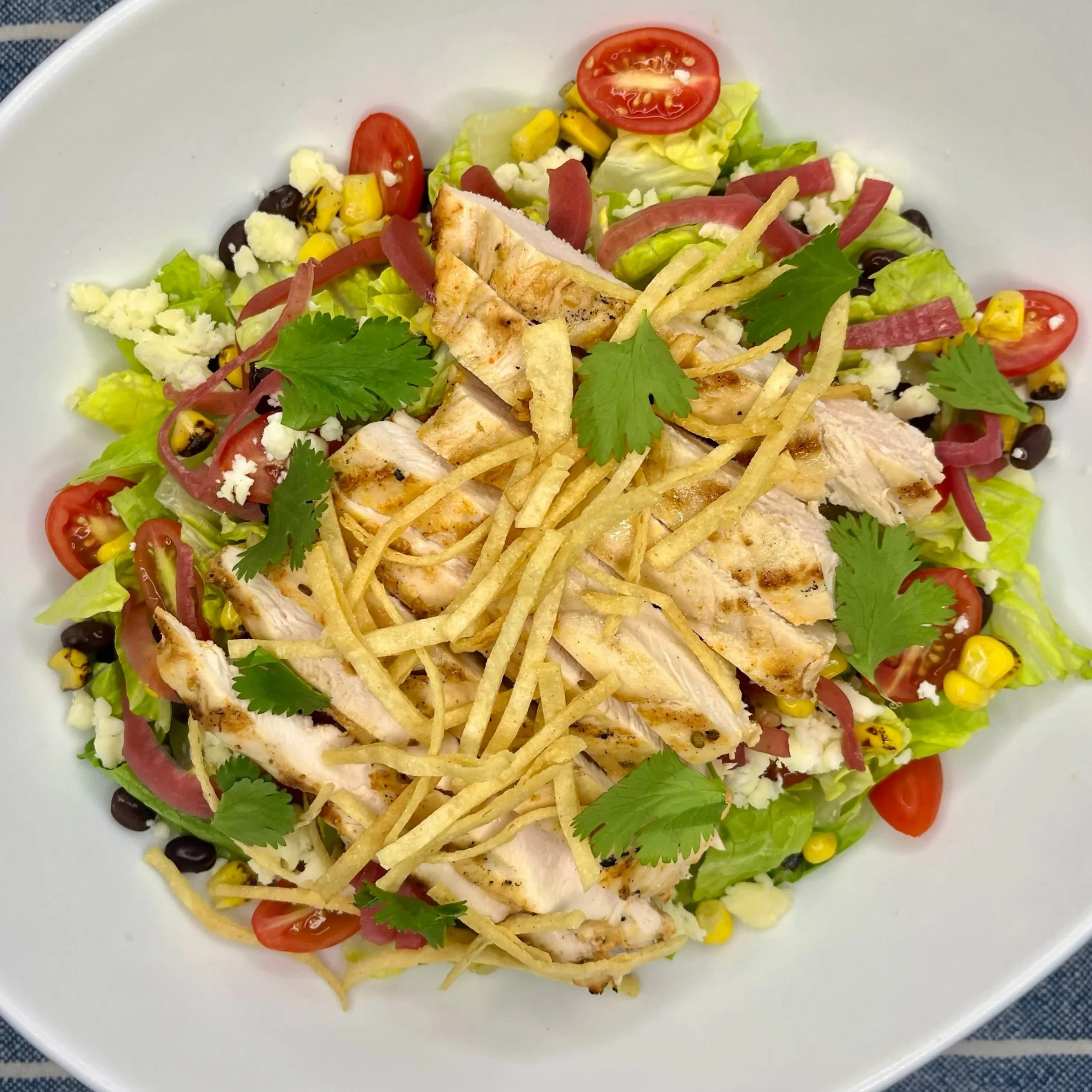 A salad with cherry tomatoes, lettuce, grilled chicken, shredded cheese, jalapenos, cilantro, thin tortilla strips, and black beans in a white bowl