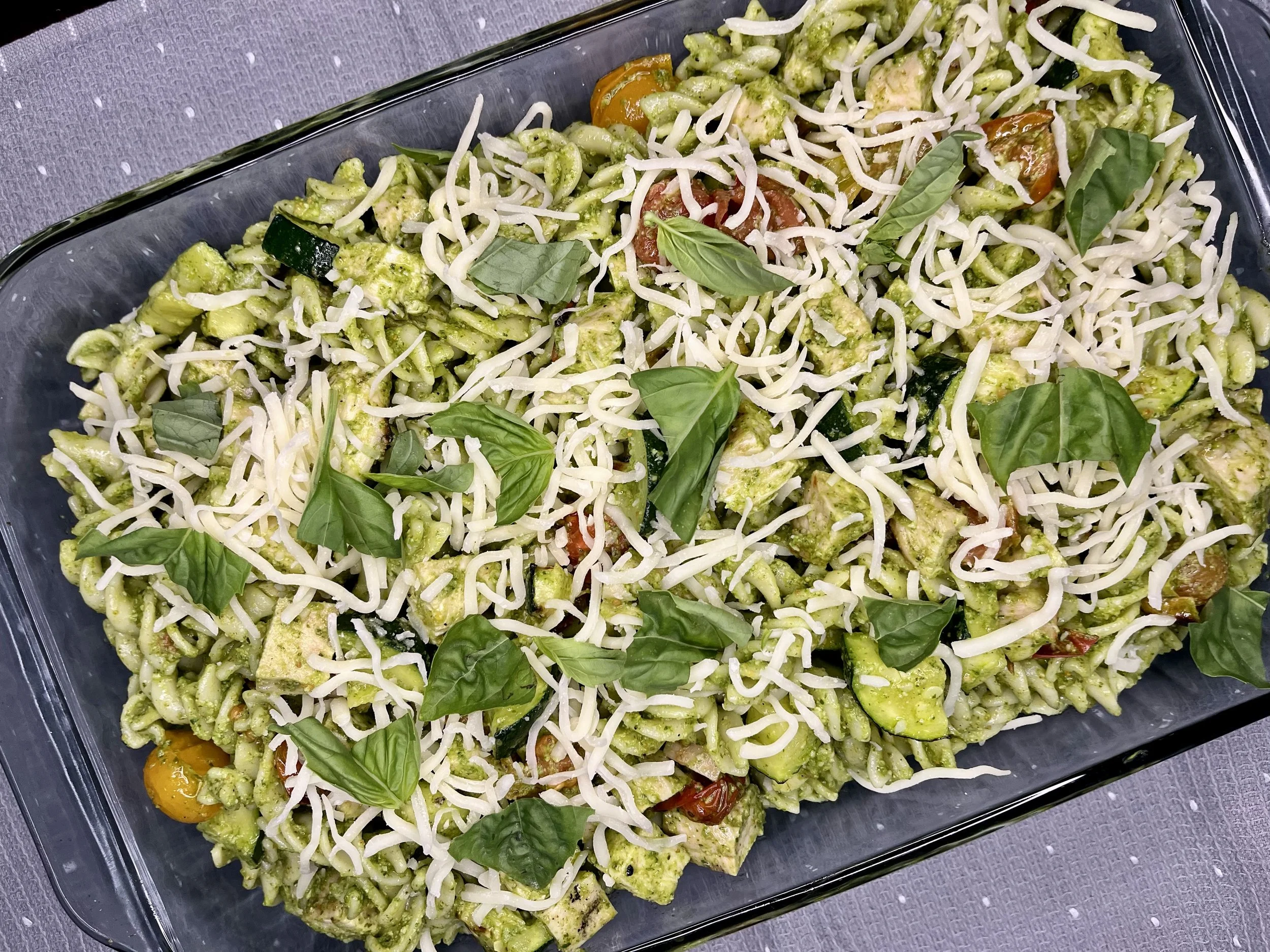A glass dish filled with caprese pasta salad garnished with fresh basil leaves. The salad contains pasta, cherry tomatoes, zucchini, and shredded cheese, coated with a green pesto dressing.