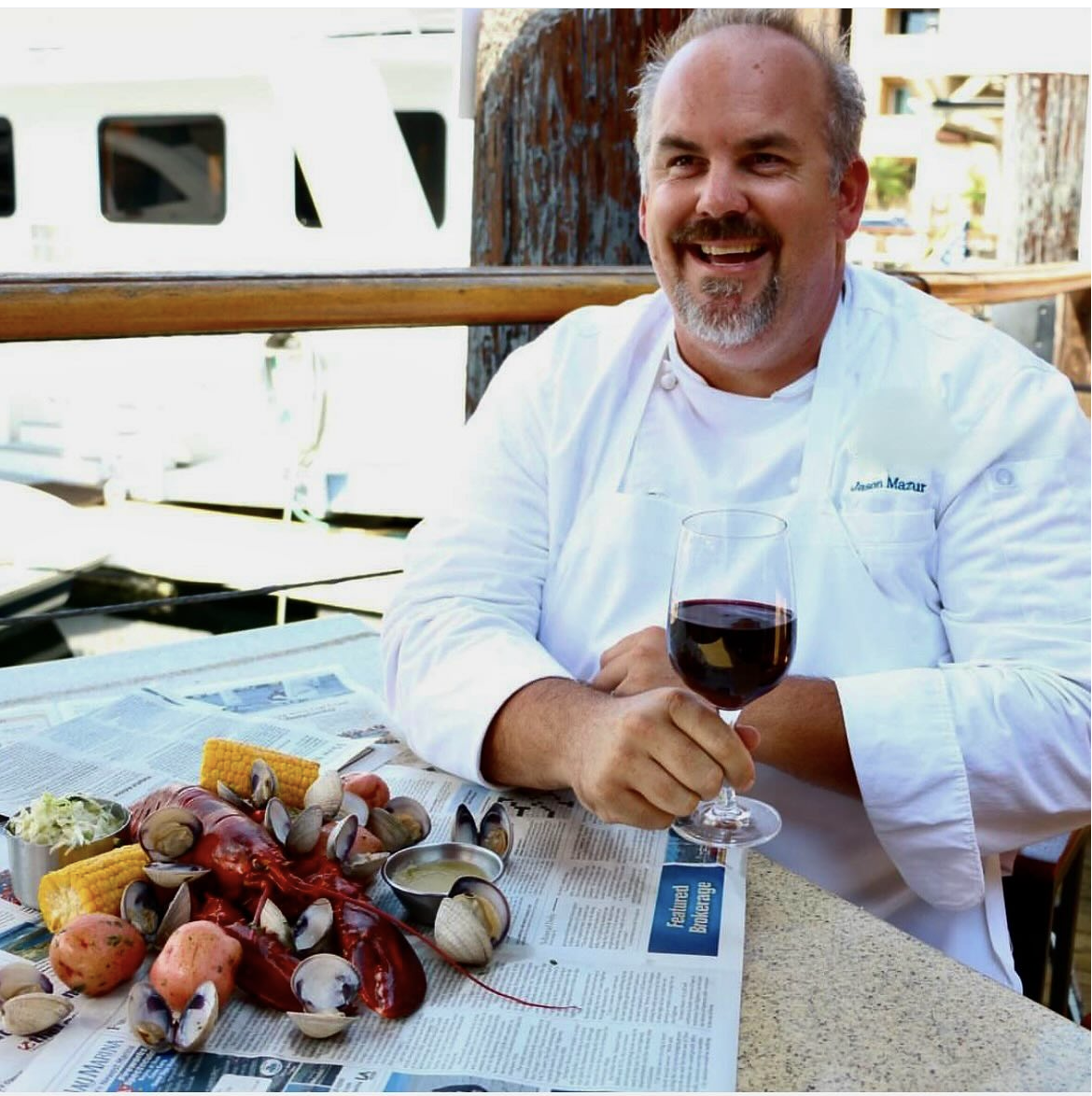 A man wearing a white chef's coat sitting at a table outdoors, holding a glass of red wine, with seafood, corn, and potatoes on the table, and smiling.