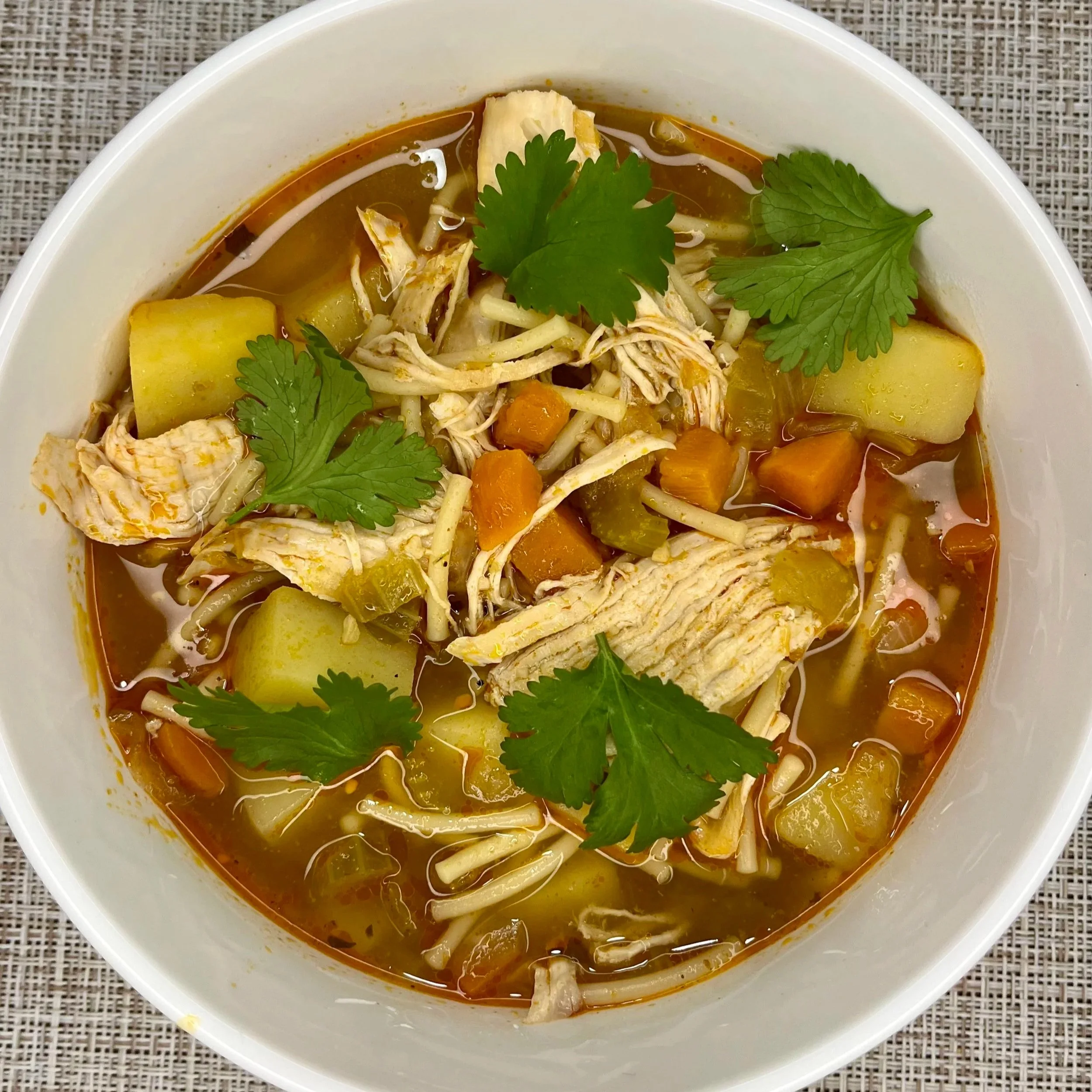 A bowl of homemade chicken soup with potatoes, carrots, shredded chicken, cilantro garnish, and spicy broth.