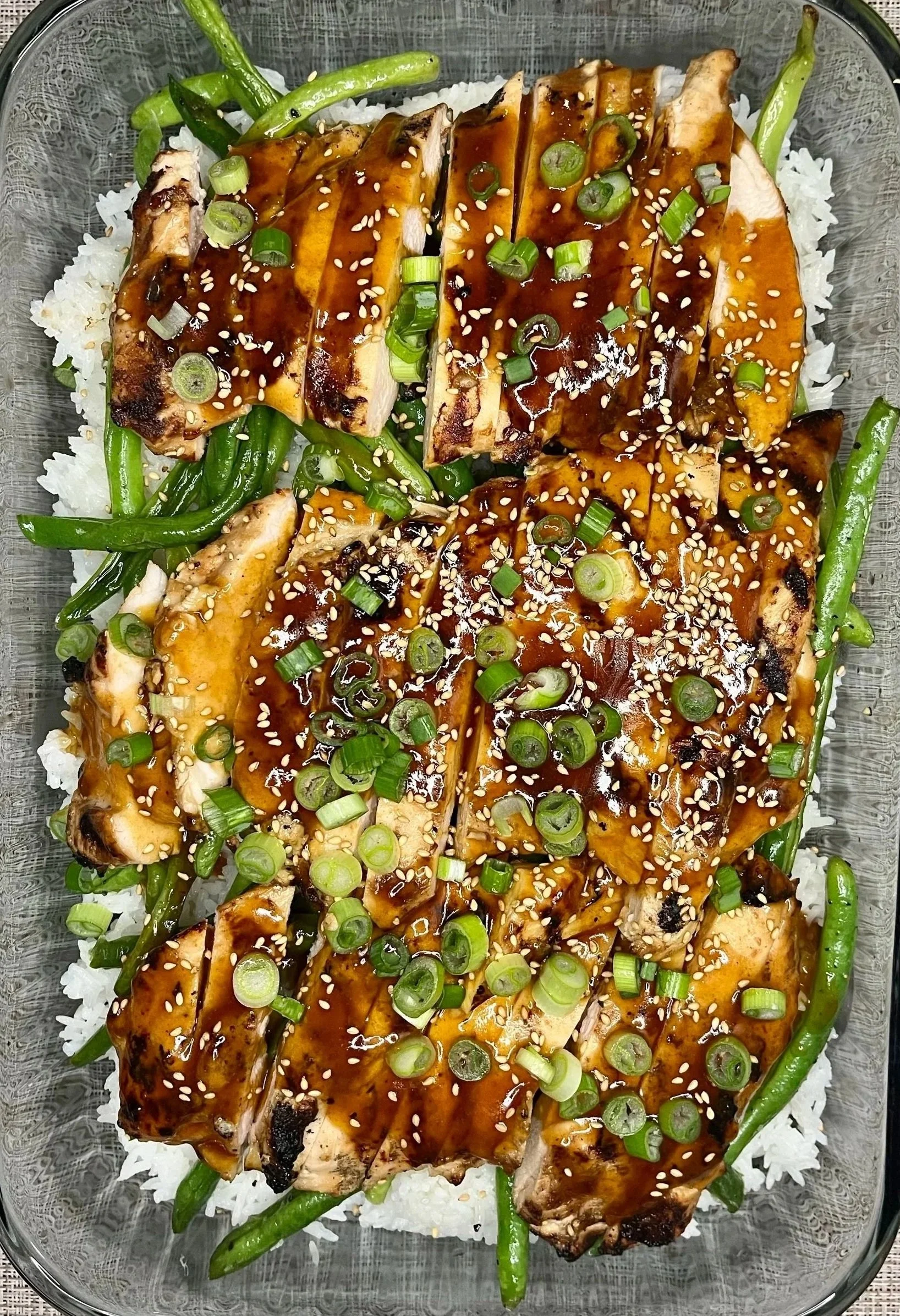 Sliced grilled chicken topped with a dark sauce and sprinkled with sesame seeds and chopped green onions, served over white rice with green beans.