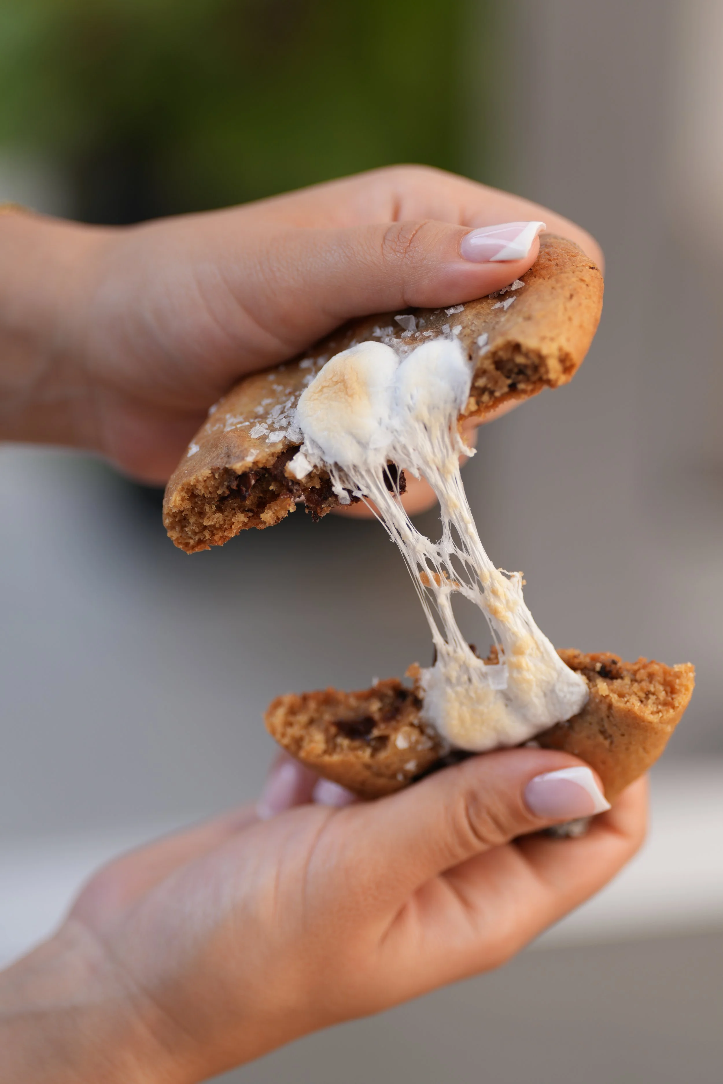 Close-up of hands holding a chocolate chip cookie with melted cheese stretching between halves.