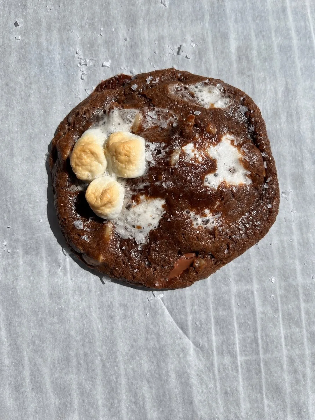 Close-up of a chocolate cookie topped with white marshmallows and chocolate chunks on a silver surface.