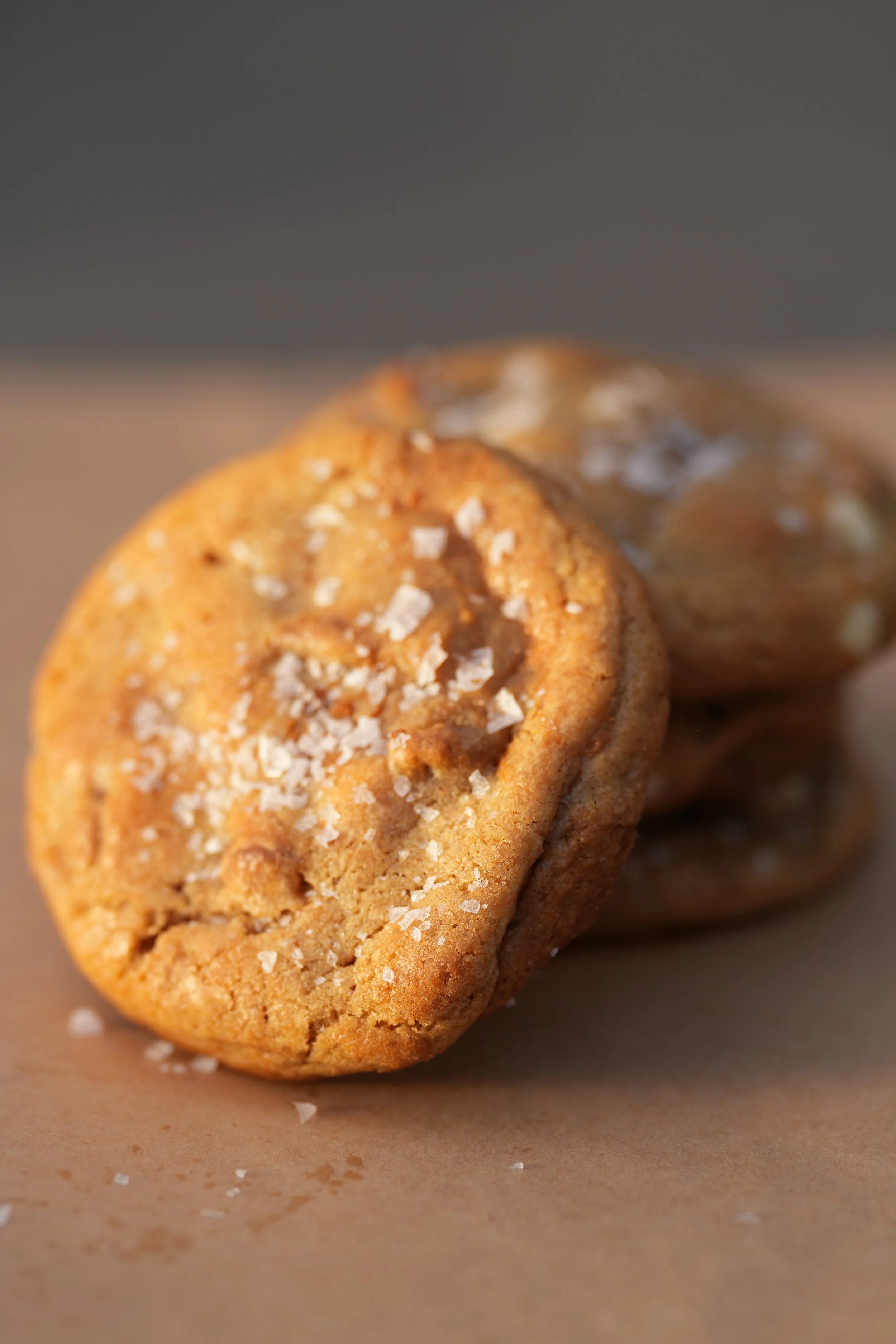 Close-up of a stack of three salted caramel cookies with coarse salt on top, on a brown paper surface.