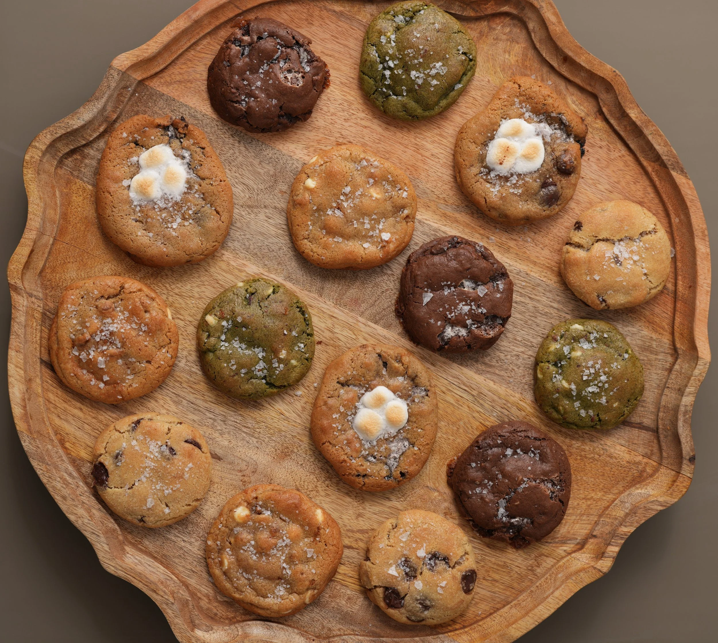 Assorted cookies with salt flakes on a wooden platter, some topped with marshmallows and chocolate chips.
