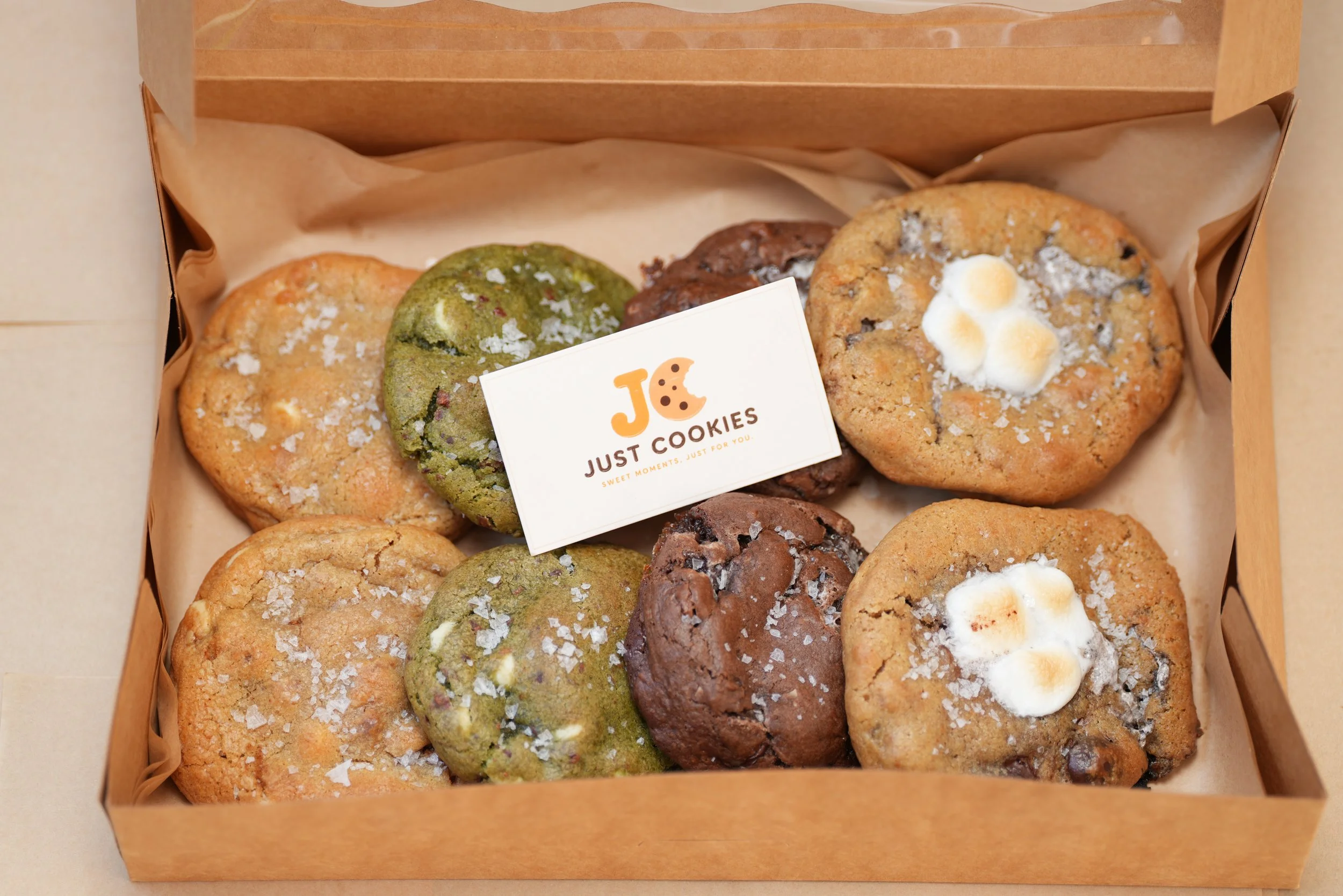 A box of assorted cookies, including green matcha, chocolate chip, and oatmeal with marshmallows, with a label that reads 'Just Cookies' and 'Sweet moments, just for you'.