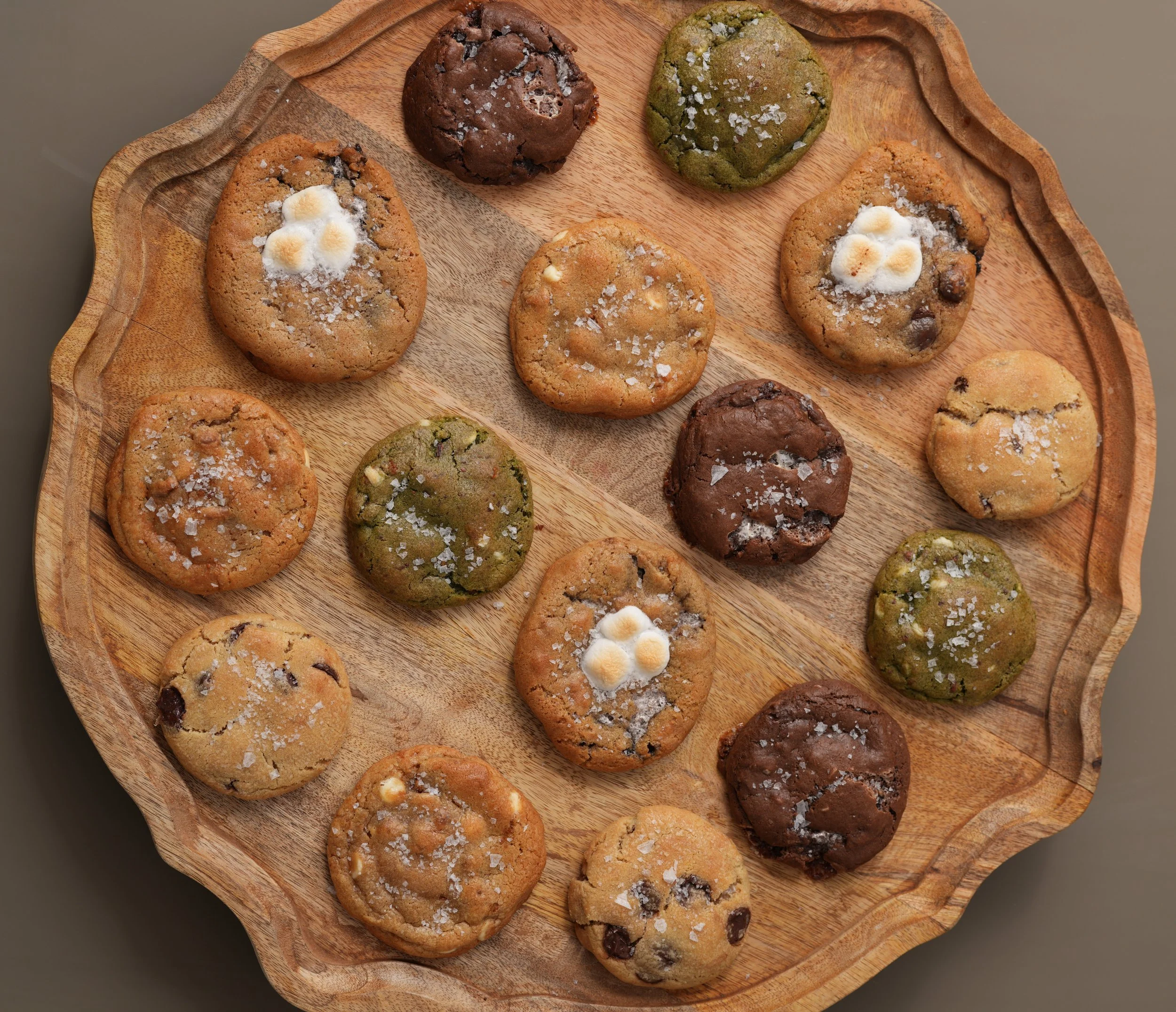 Assorted cookies on a round wooden tray, including chocolate chip, green matcha, and oatmeal with white chocolate chips, sprinkled with coarse salt.