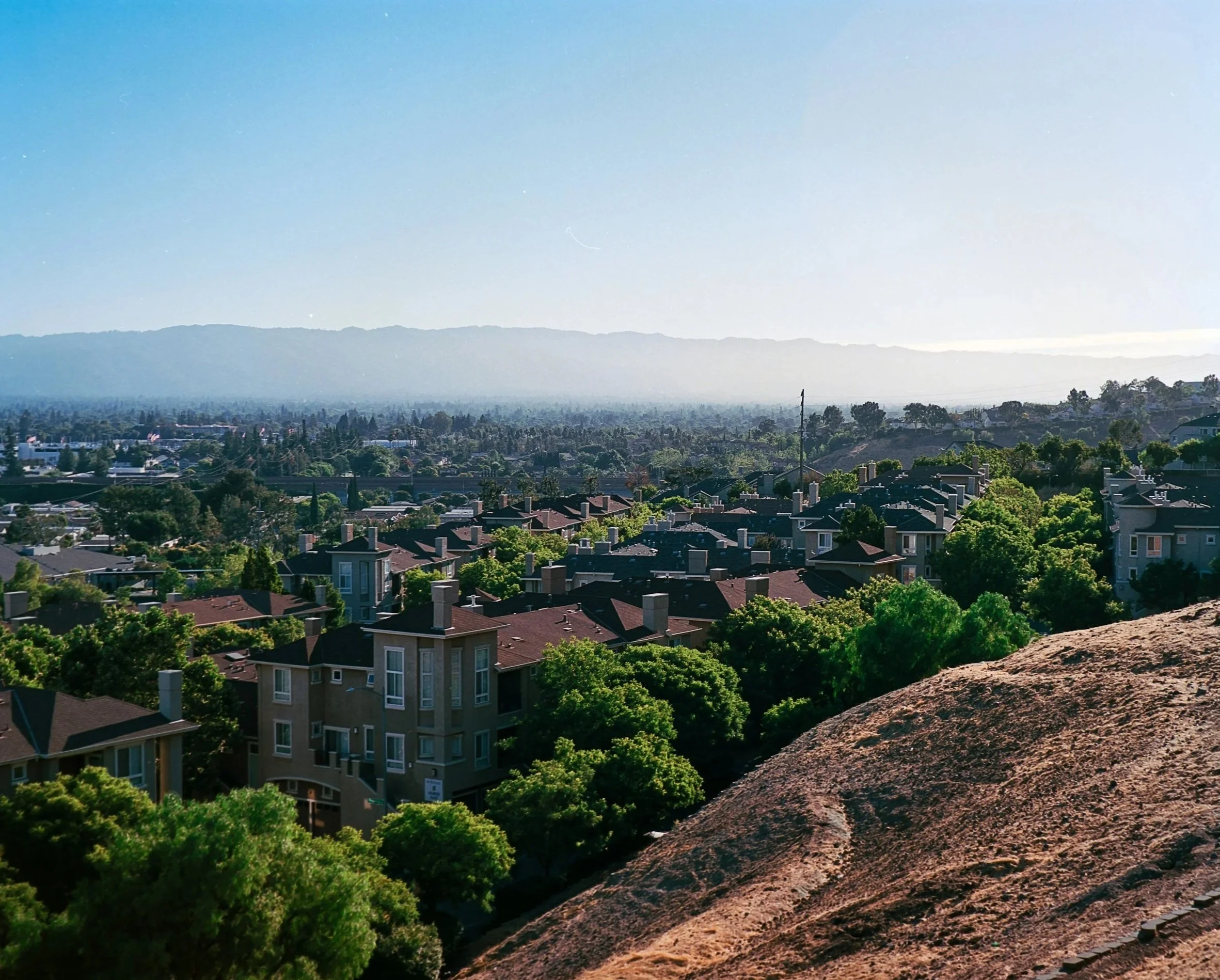 View of a suburban neighborhood with multiple houses and trees, with mountains in the background under a clear blue sky.