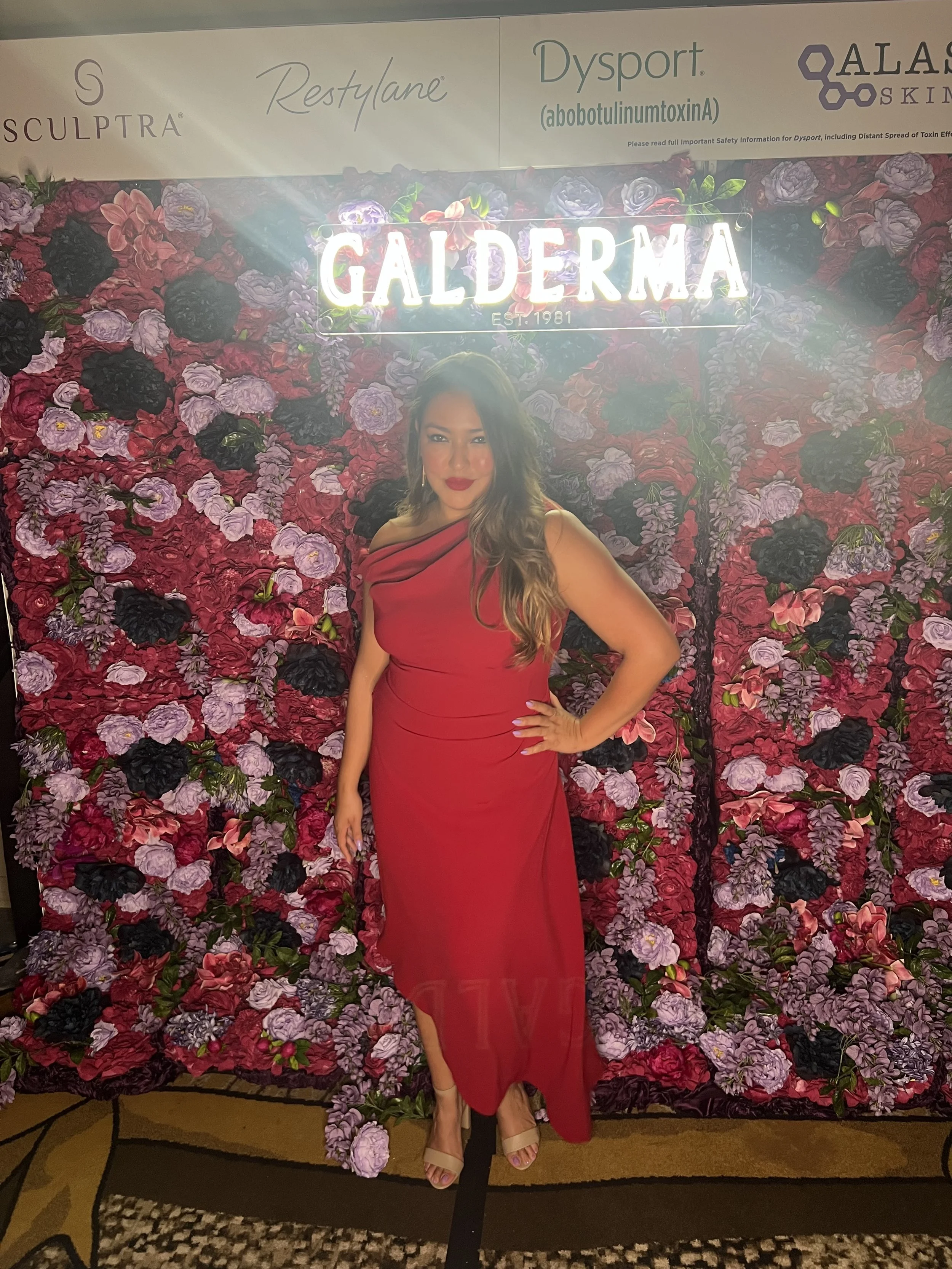 A woman in a red dress standing in front of a floral backdrop with a neon sign reading 'GALLERMA' and the establishment date 'EST. 1981'.