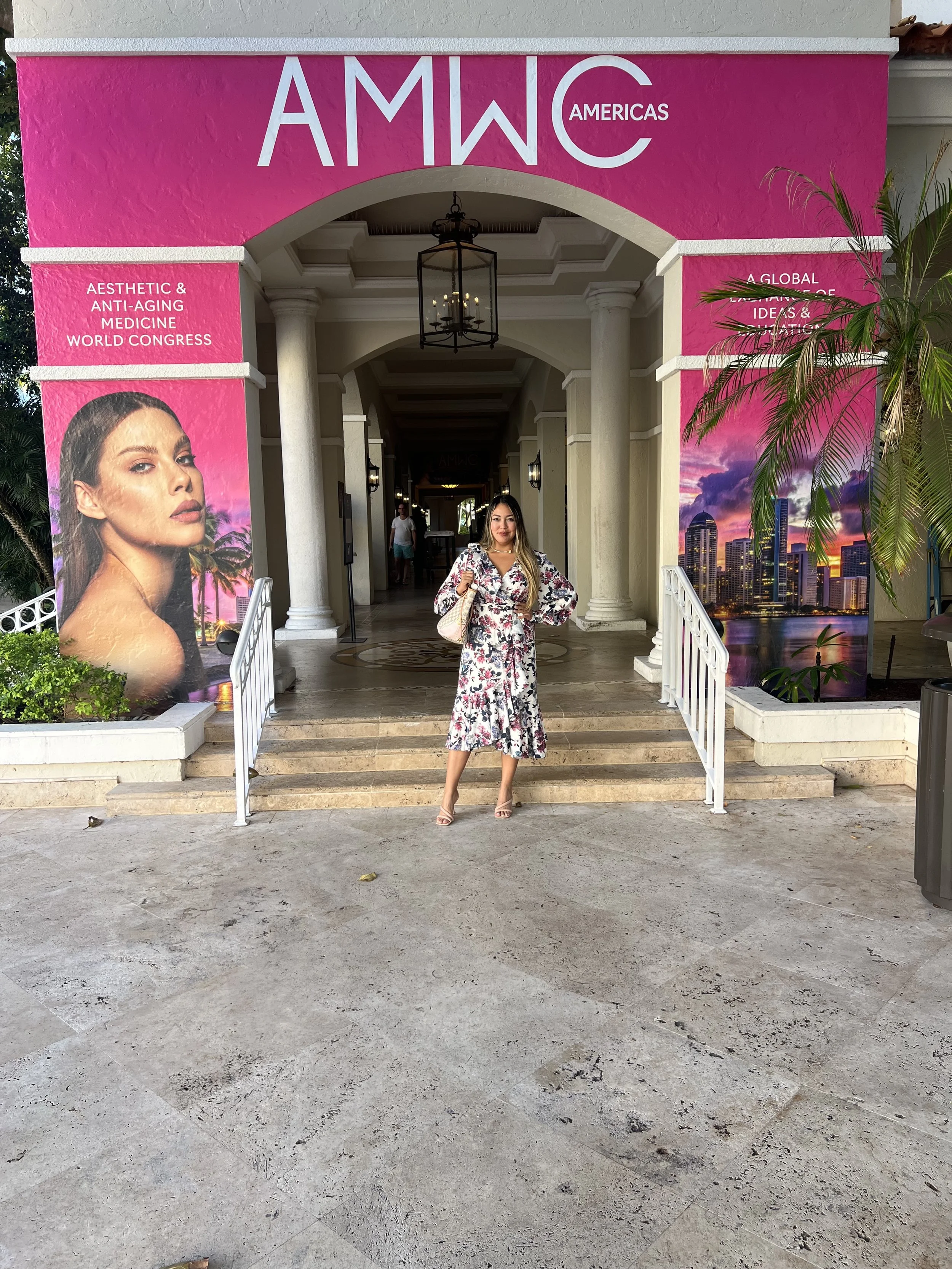 A woman in a floral dress standing at the entrance of a building, posing for a photo. The building has pink banners on either side that advertise the AMWC Americas conference, focusing on aesthetic and anti-aging medicine, with images of cityscapes a